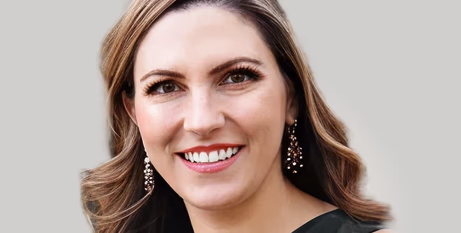 Smiling woman with wavy brown hair wearing dangling earrings, photographed against a light neutral background.