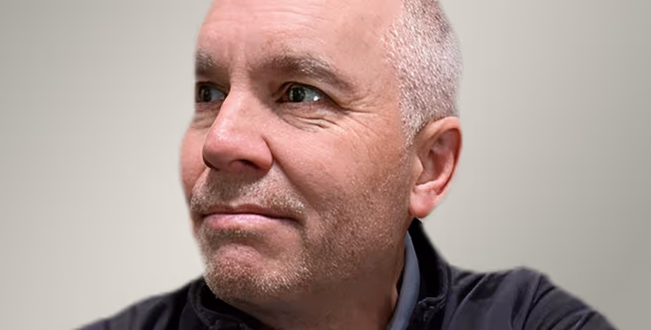 Close-up of a middle-aged man with short gray hair and stubble, looking to the side against a neutral background.