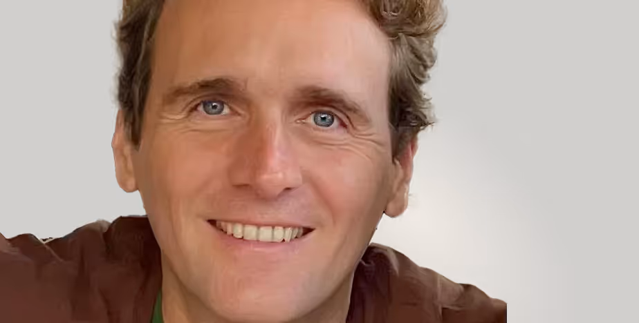 Man with short curly hair and blue eyes smiling warmly at the camera against a soft gray background.