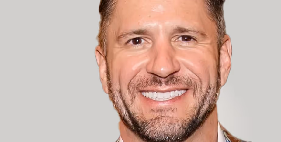 Bearded man smiling in a close-up portrait, wearing a suit jacket and standing against a light neutral backdrop.
