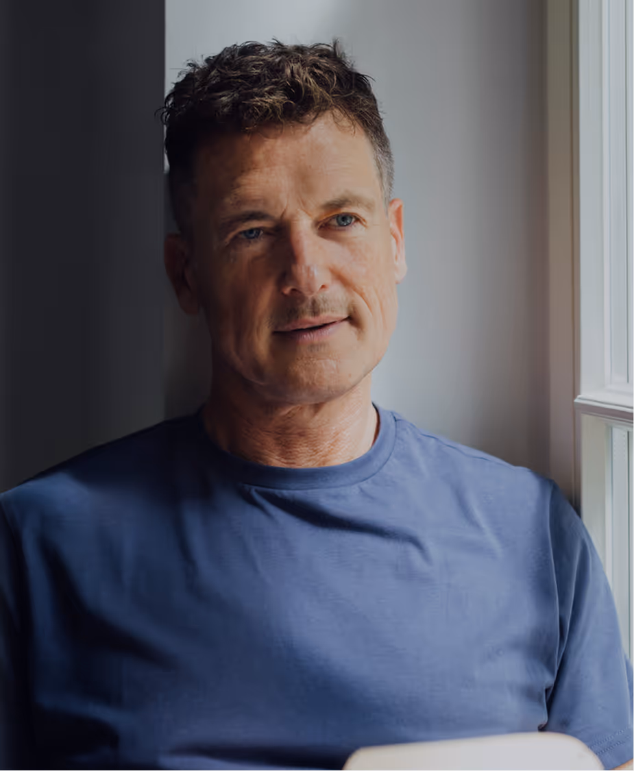 Man sitting by a window in soft natural light, wearing a blue shirt and looking thoughtful.