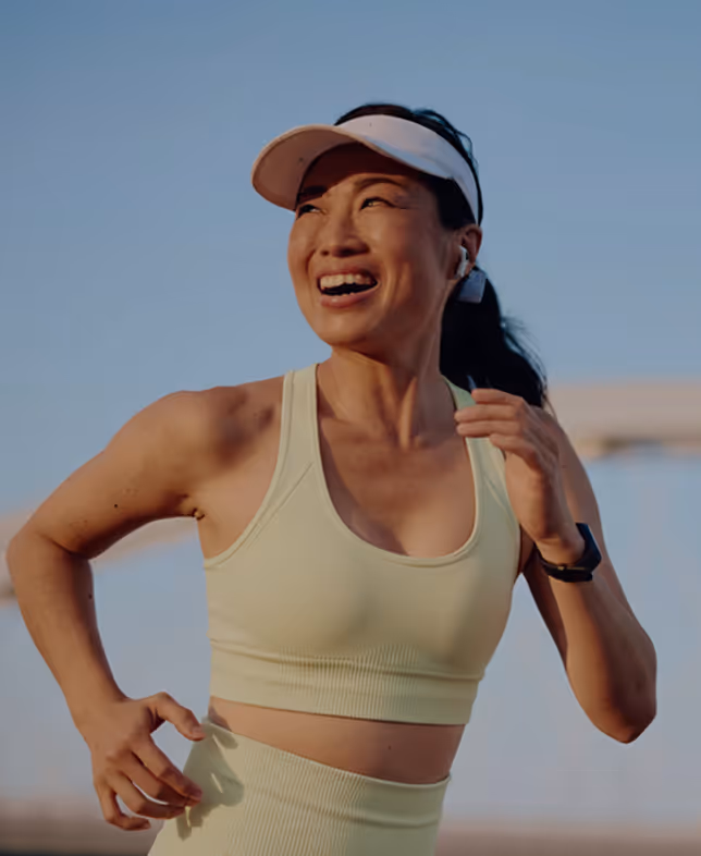 Woman running outdoors at sunrise, smiling while wearing a light yellow sports outfit, visor, and wireless earbuds.