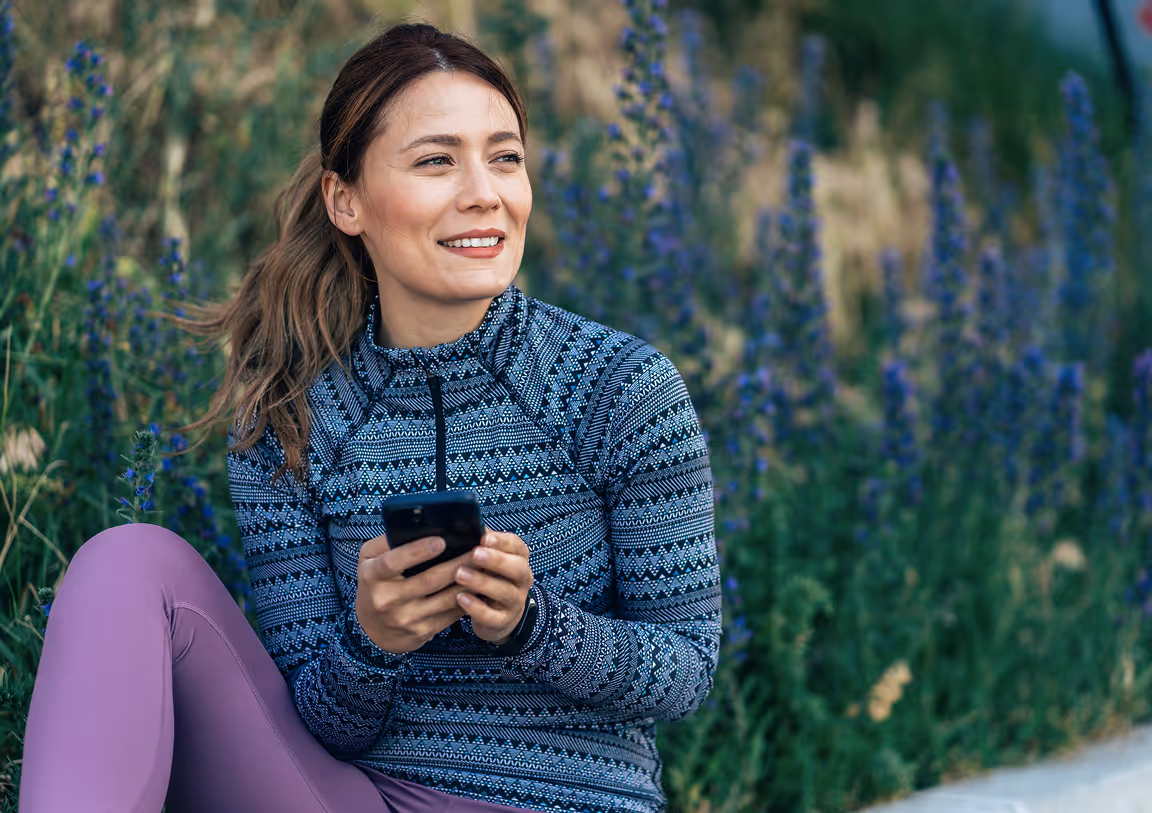 Woman sitting outdoors by wildflowers, wearing athletic clothing and holding a smartphone while smiling.