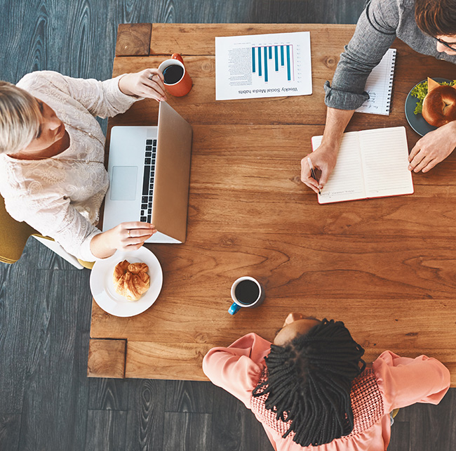 Overhead view of three people at a wooden table with coffee, pastries, a laptop, a notebook, and a printed chart.