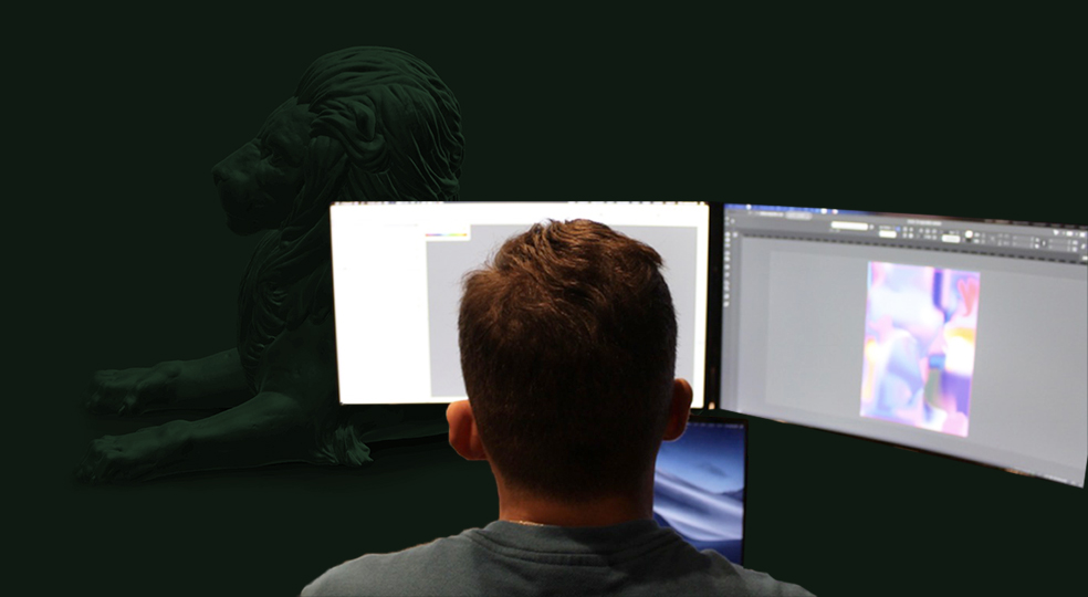 Person from behind working on a computer with two large monitors displaying graphic design software, with a dark lion statue in the background.
