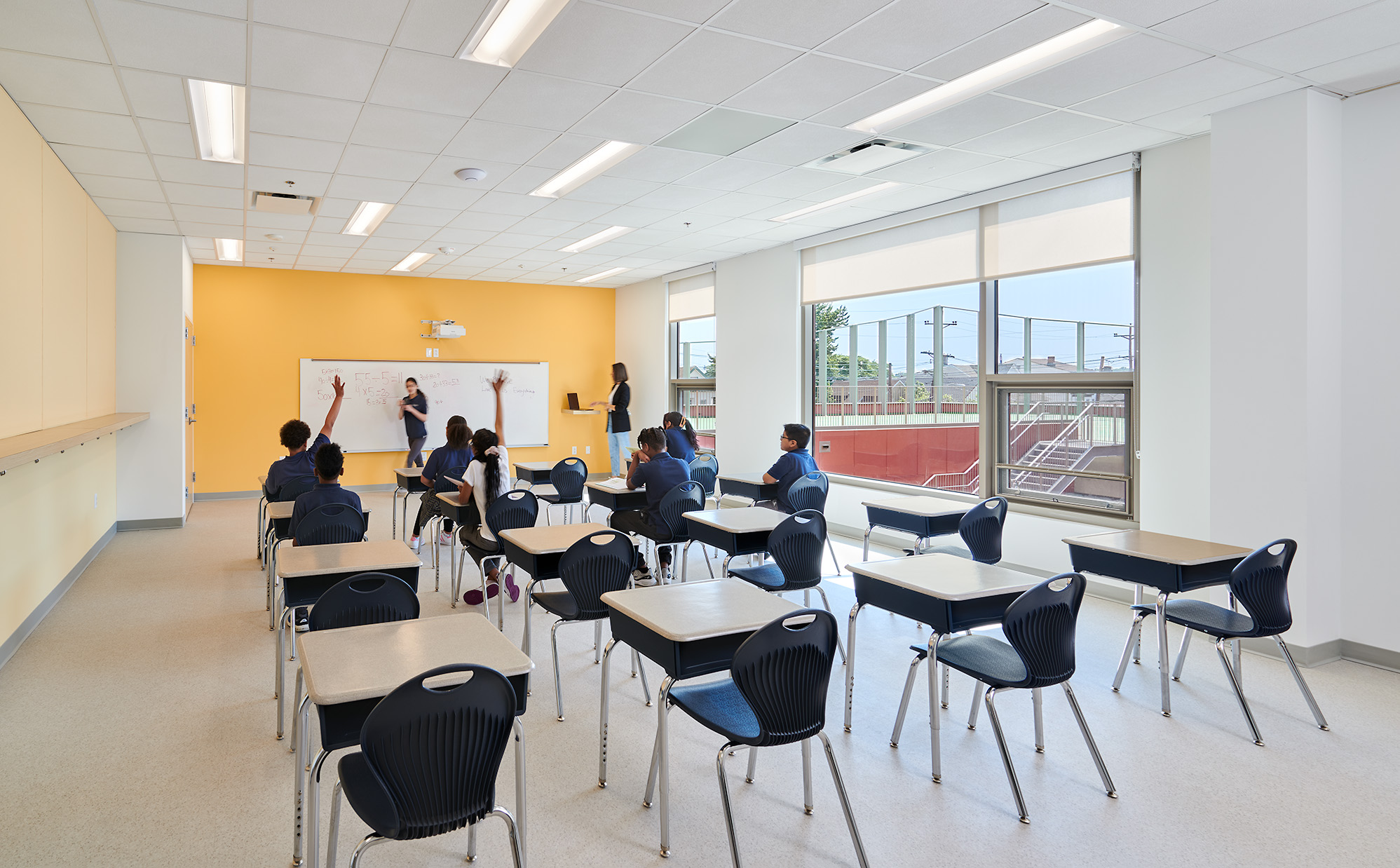 Classroom with students seated at desks facing a teacher and a whiteboard on a yellow wall, with large windows letting in natural light.