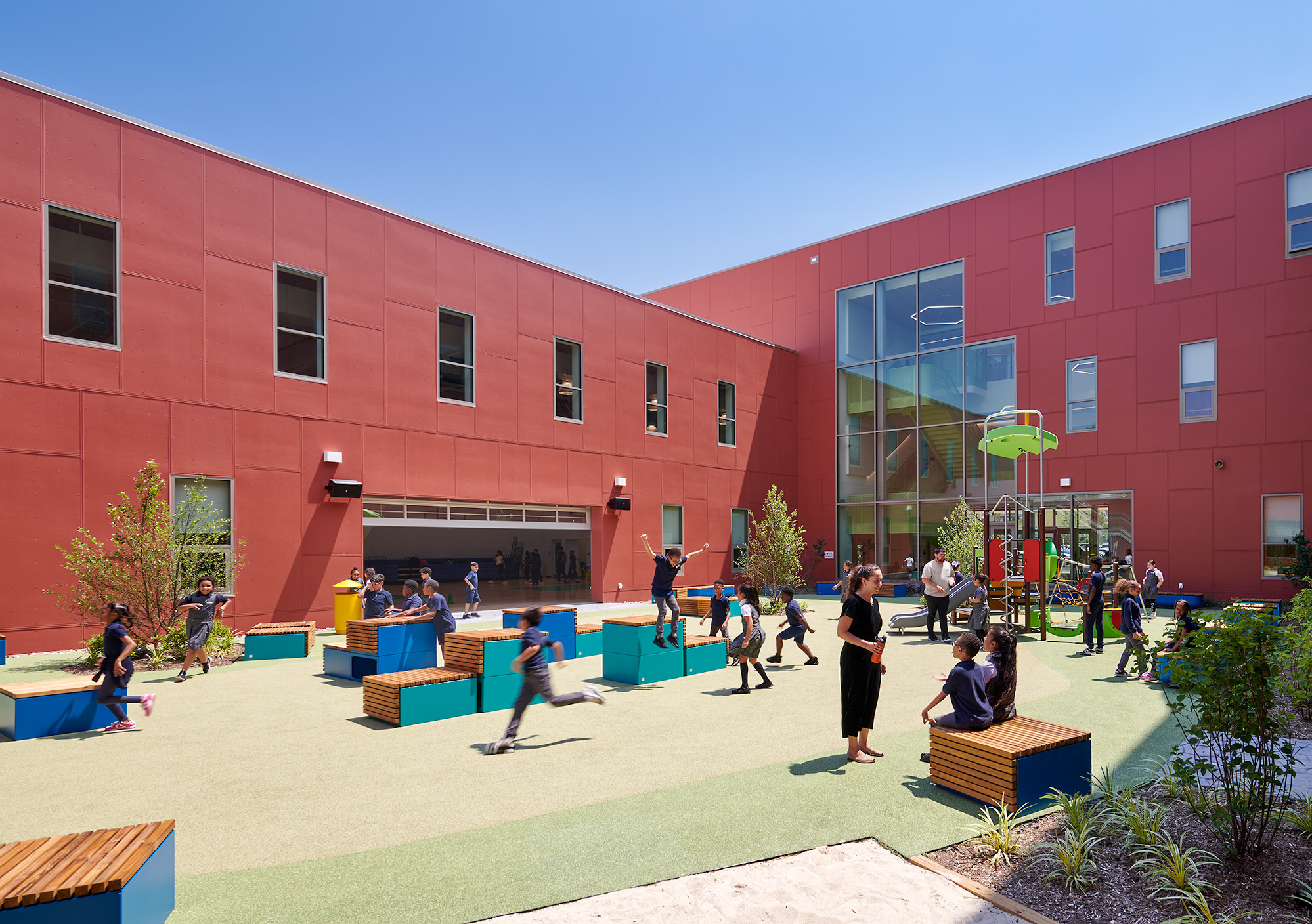 Children playing and interacting in a spacious school courtyard with red buildings, benches, and playground equipment under a clear blue sky.