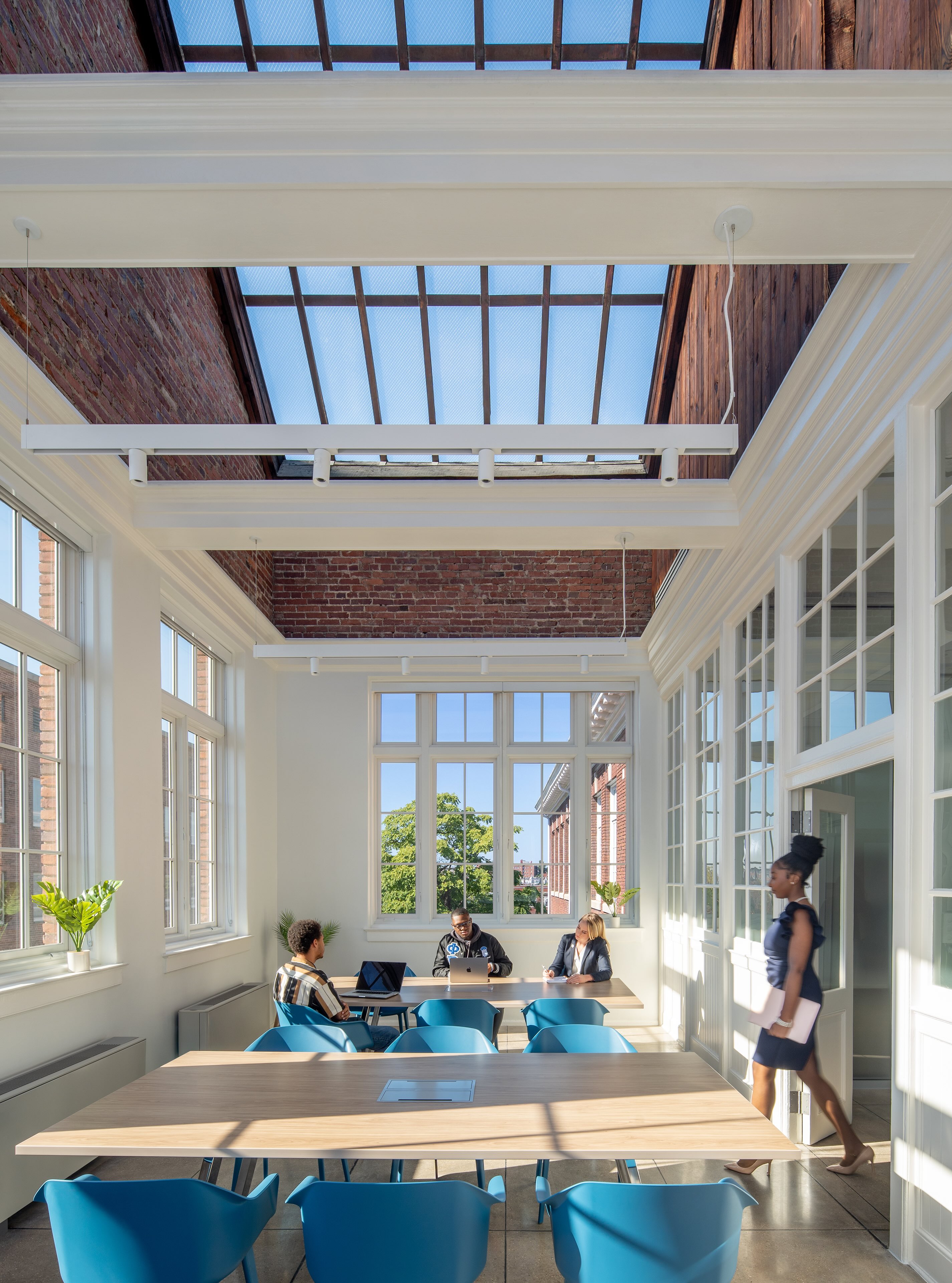 Bright conference room with skylight, blue chairs, wooden tables, and three people seated while a woman walks by holding documents.