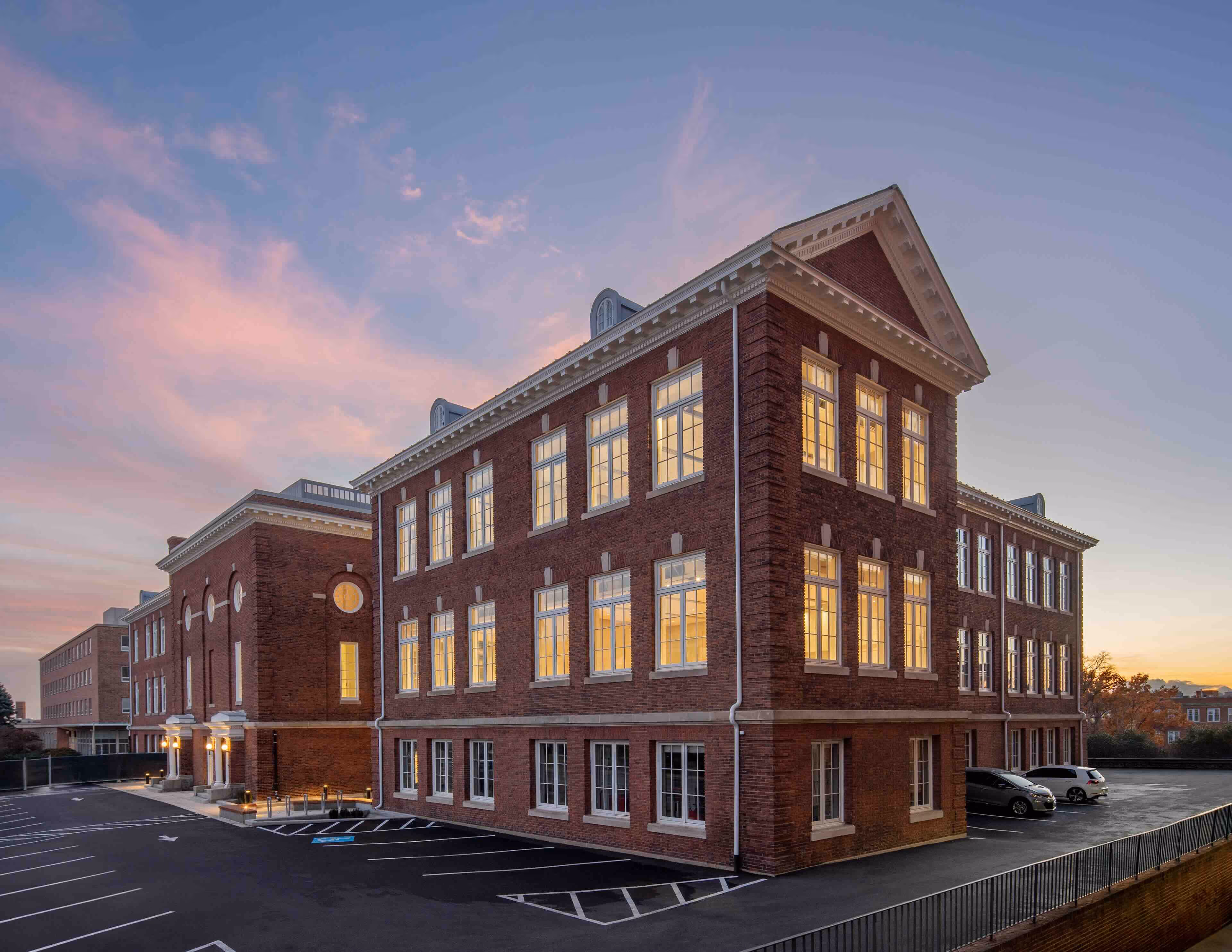 A large, illuminated brick building with multiple windows at dusk and an empty parking lot in front.