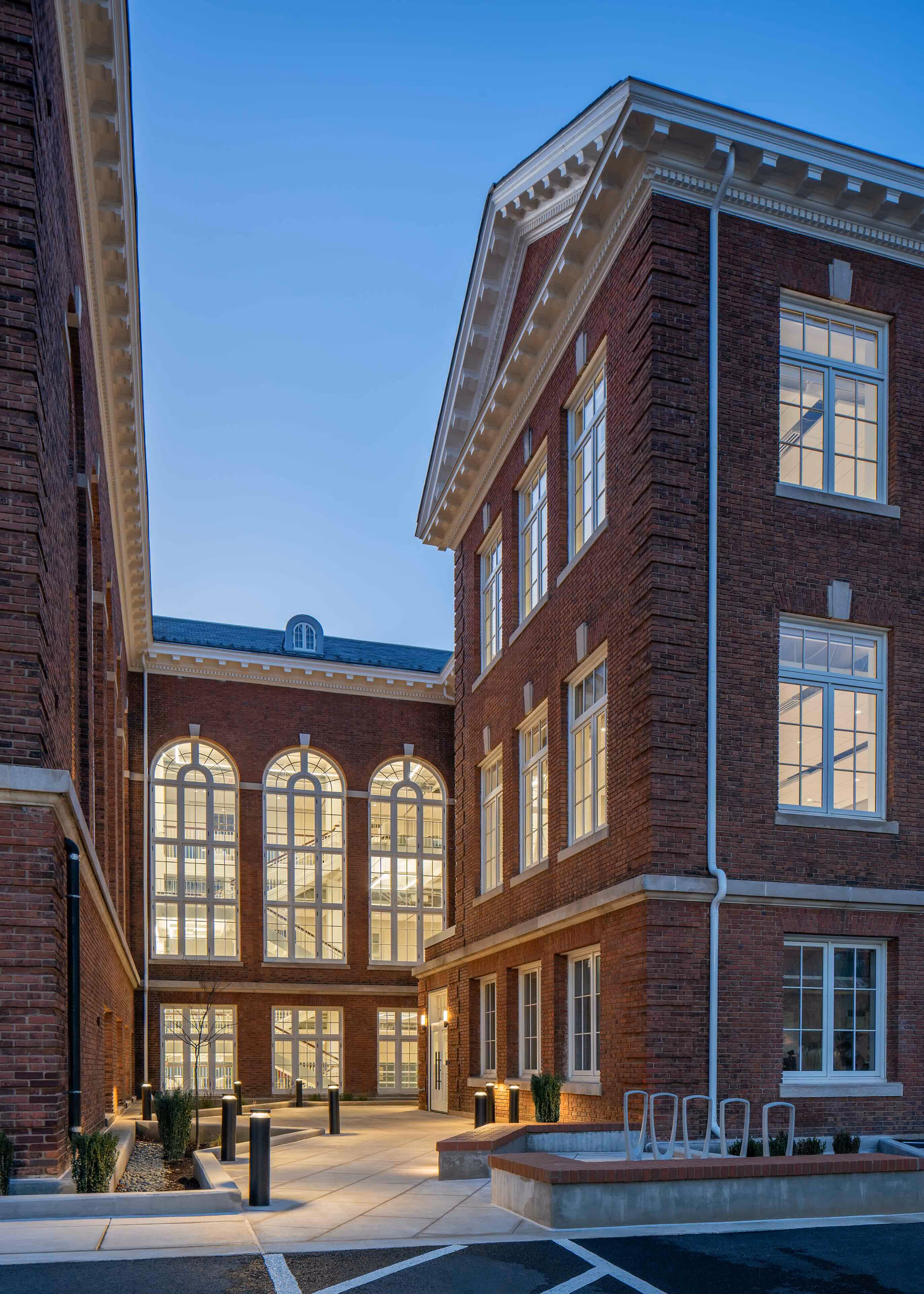 Brick building with large arched windows lit from inside at dusk with modern outdoor lighting along a paved courtyard.