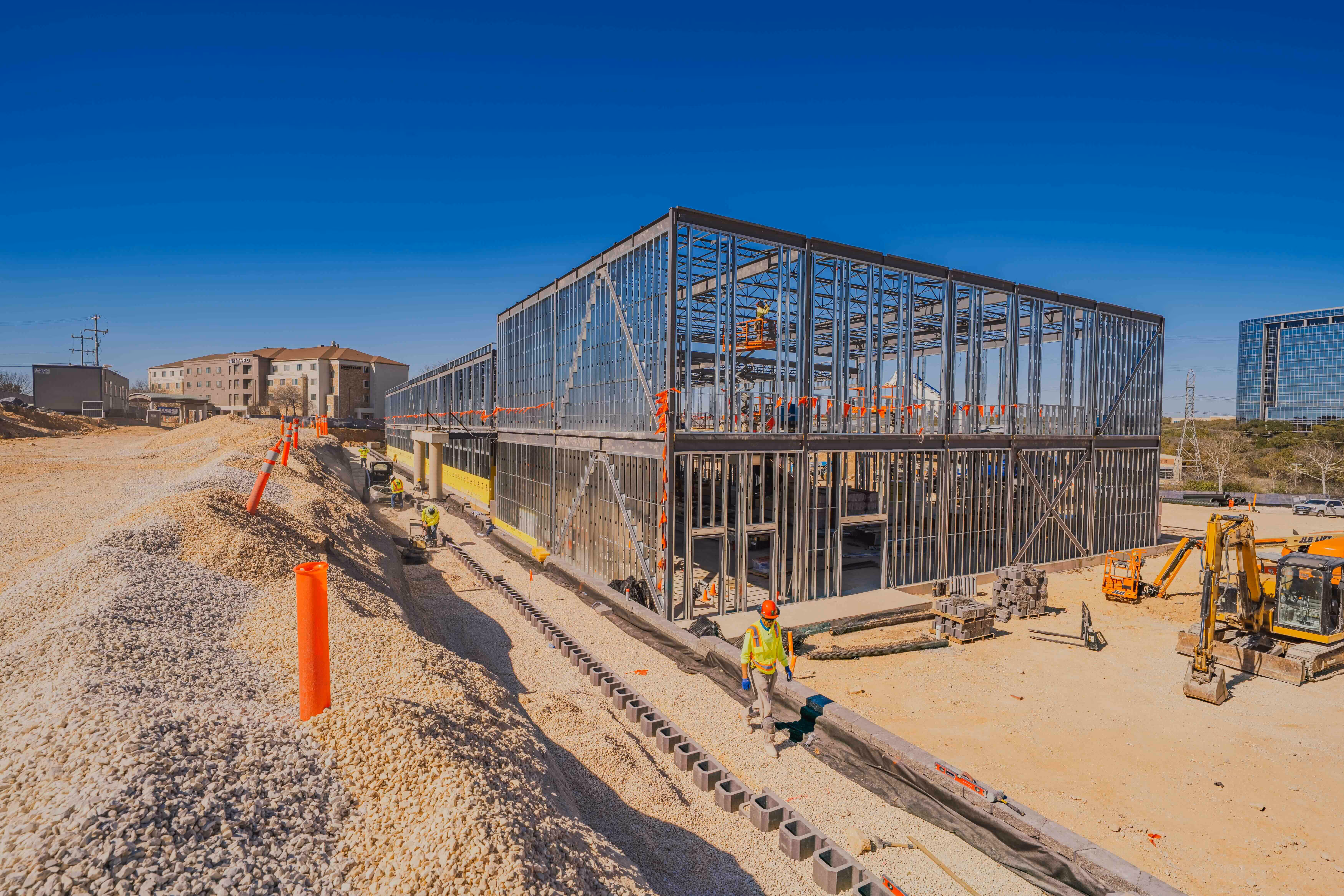 Construction site with workers building a large steel-framed commercial building under a clear blue sky.
