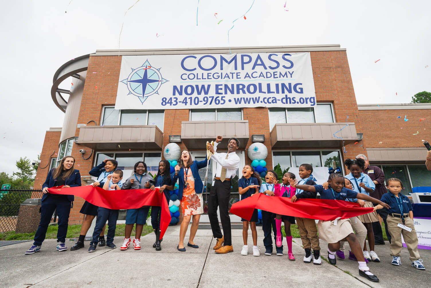 Group of joyful children and two adults holding a red ribbon and large scissors during a ribbon-cutting event in front of Compass Collegiate Academy, which has a banner announcing enrollment.