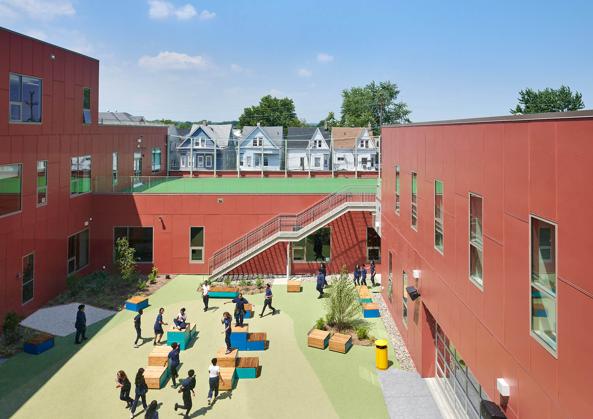 School courtyard with students walking and sitting on wooden benches amid modern red buildings under a clear blue sky.