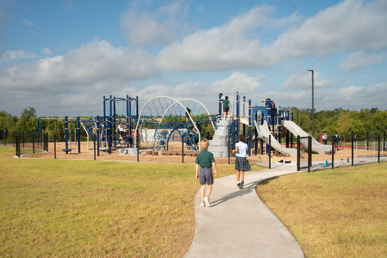 Children walking on a concrete path toward a fenced playground with climbing structures and slides under a partly cloudy sky.