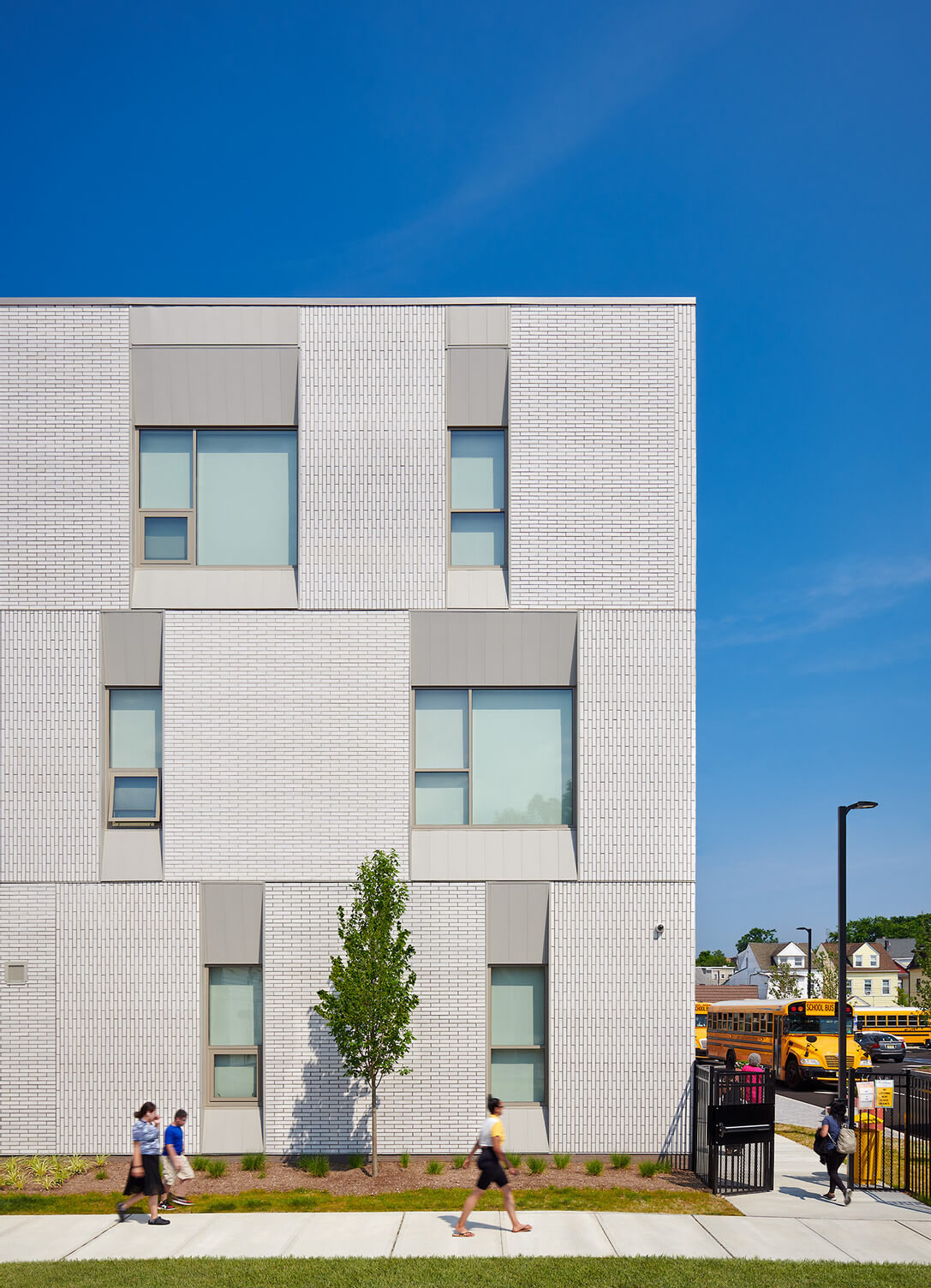 Modern white brick school building with large windows, a small tree, people walking on the sidewalk, and yellow school buses in the background.