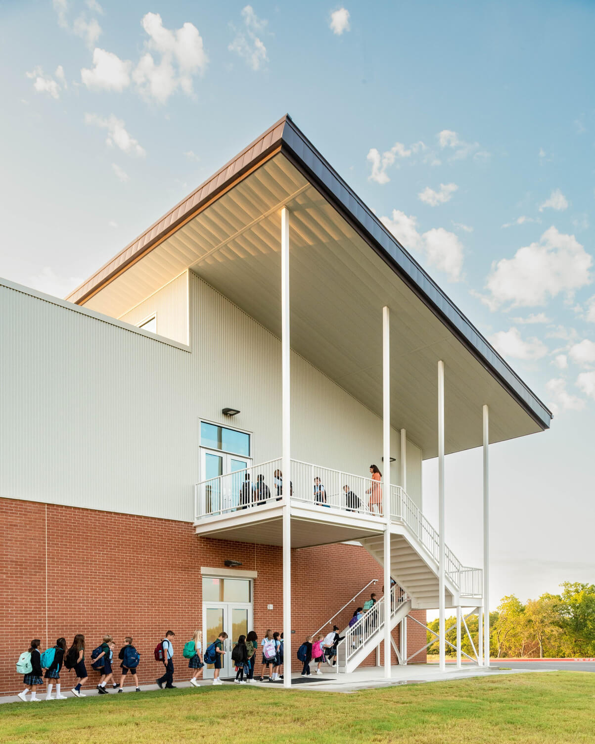 Line of young children with backpacks walking along the side of a modern school building with a staircase and a covered balcony.