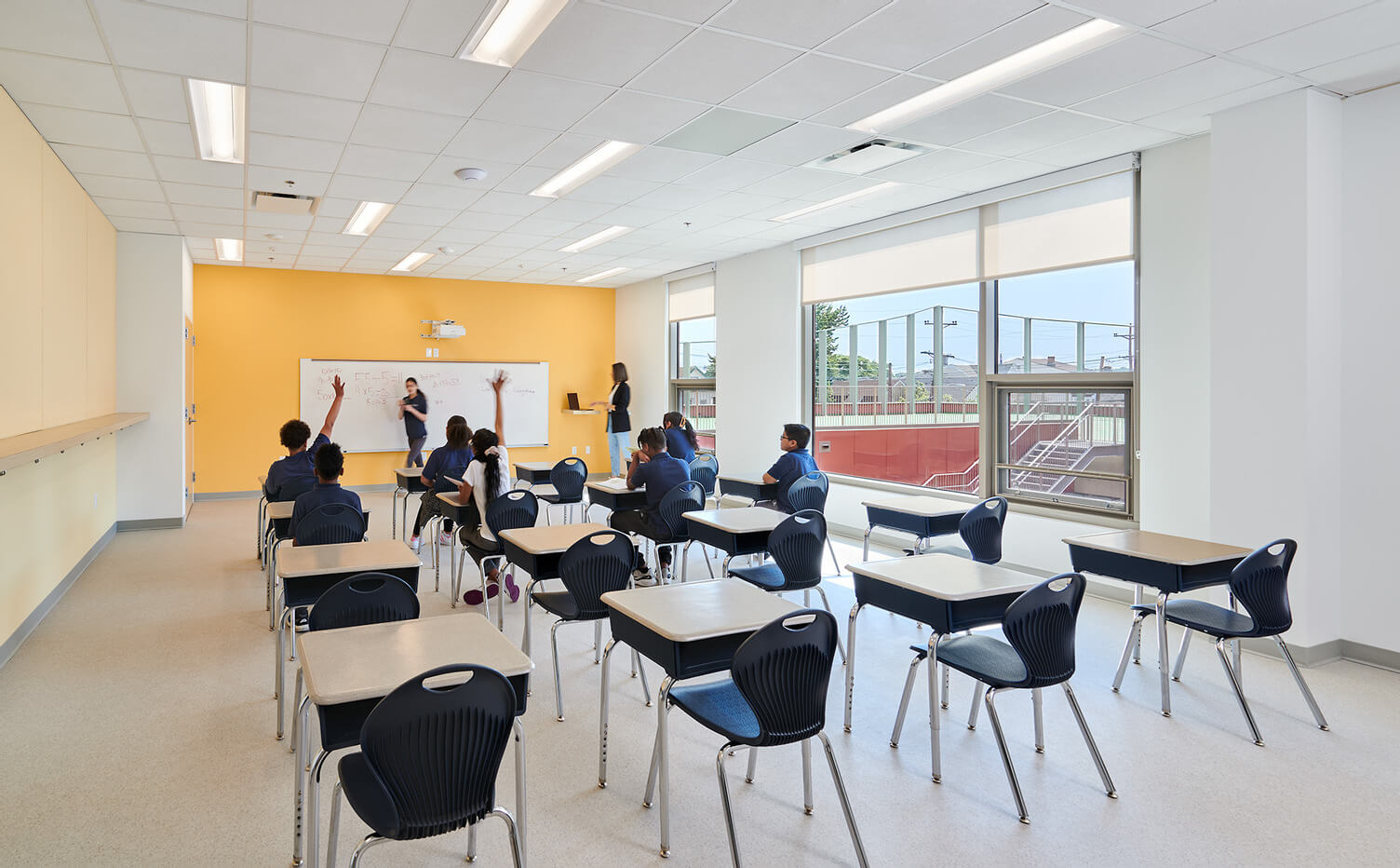 Classroom with students sitting at individual desks while a teacher writes on a whiteboard and another teacher assists.