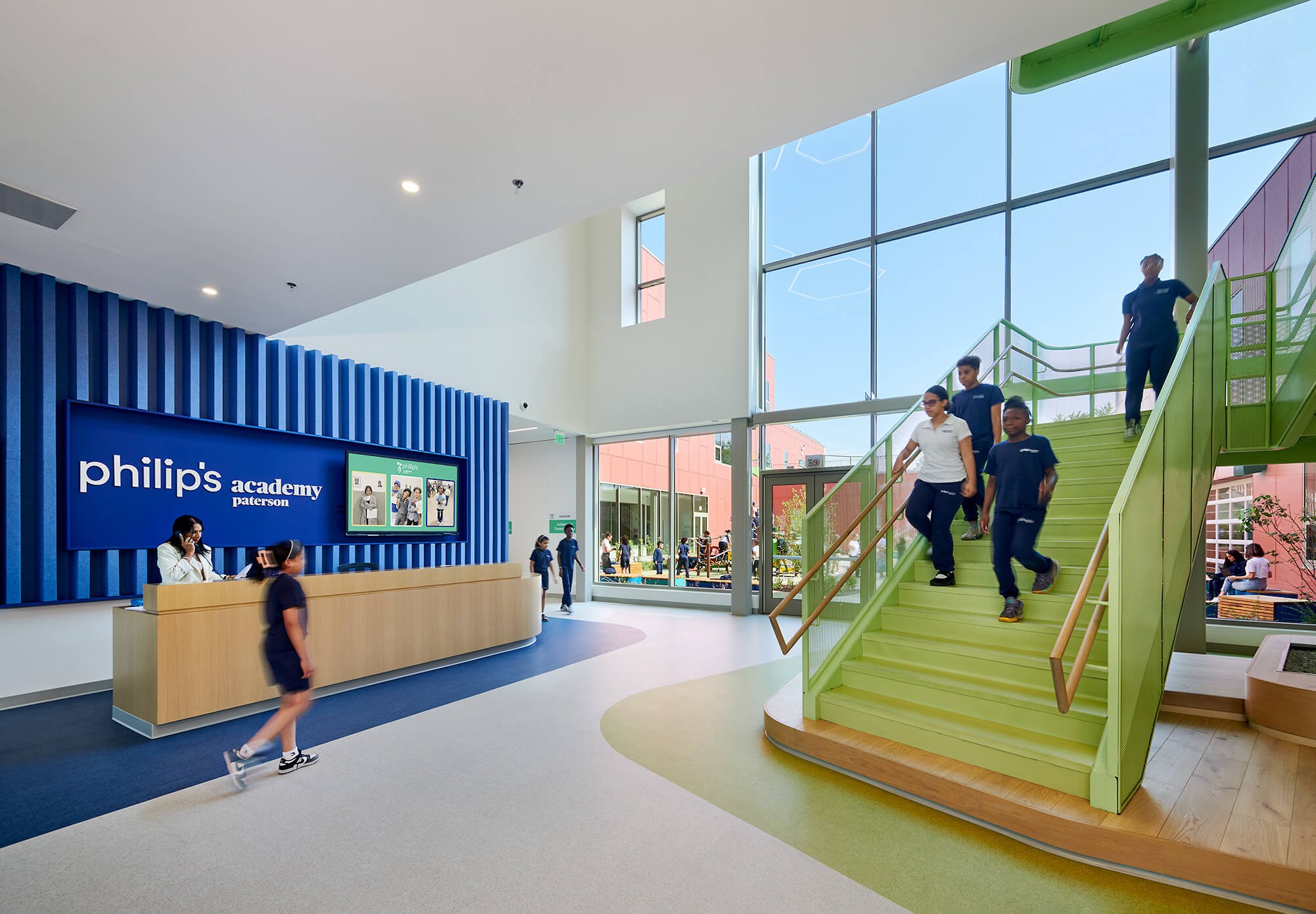 Children in navy uniforms walking down green stairs in a spacious school lobby with large windows and a reception desk under a sign reading 'philip's academy paterson'.