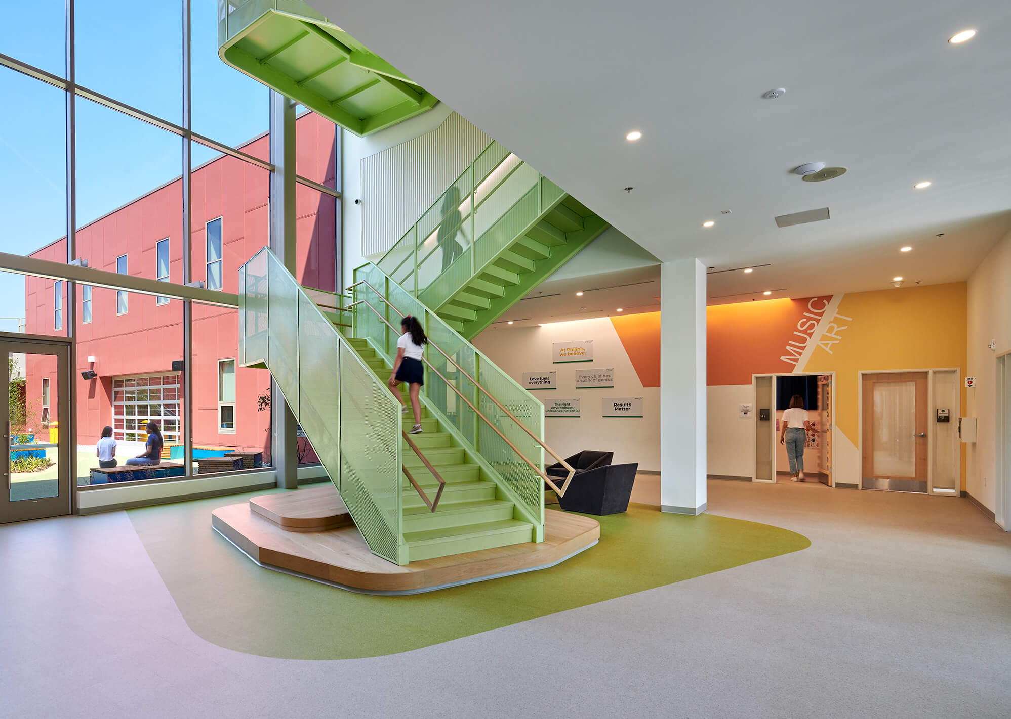 Modern indoor stairway painted light green with a woman walking up, large window showing two people sitting outside near a red building, interior walls with colorful geometric design and text, and another woman walking through a door under 'Music Art' signage.