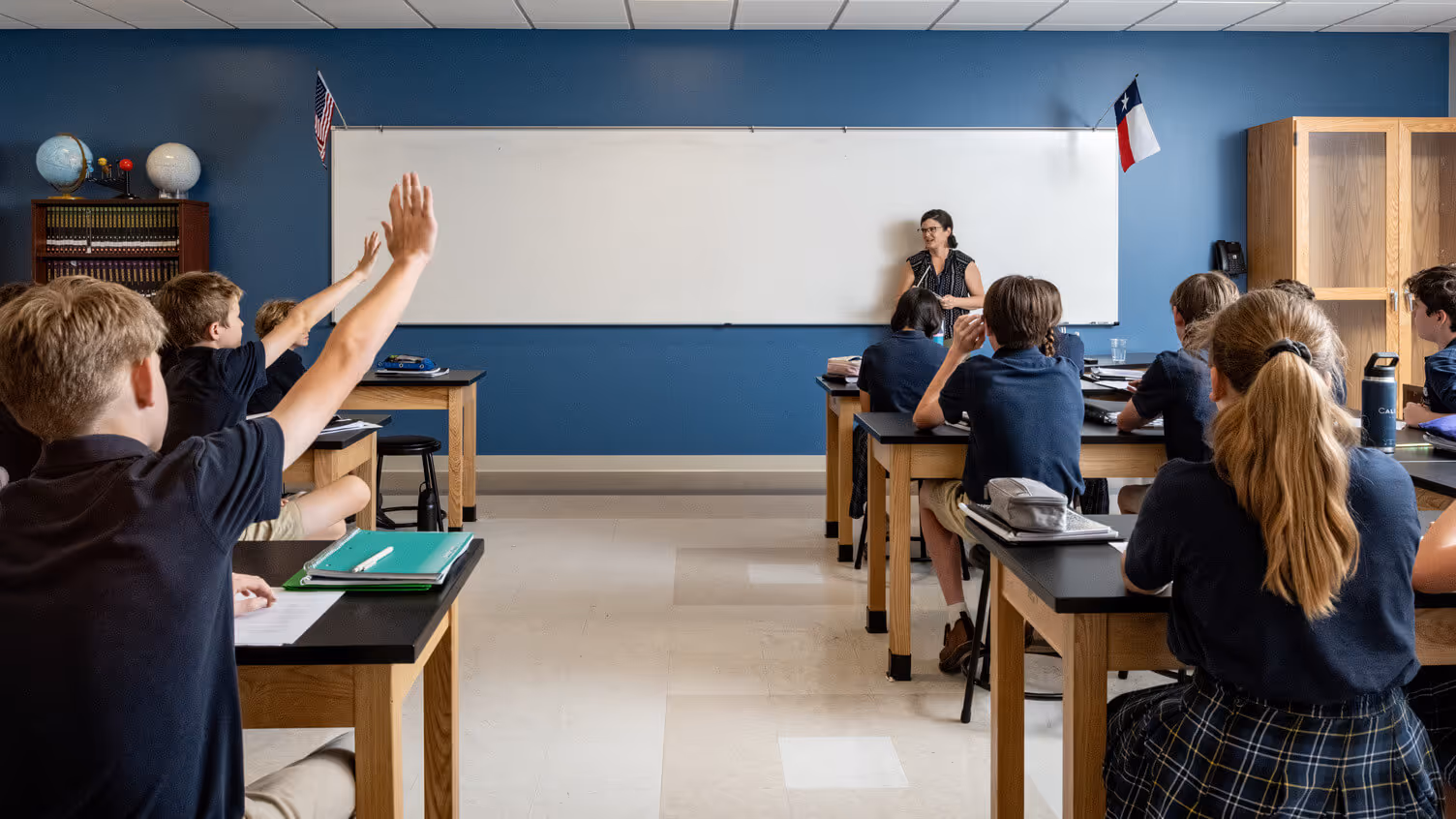 Classroom of students in uniforms sitting at desks with raised hands while a teacher stands by a whiteboard.