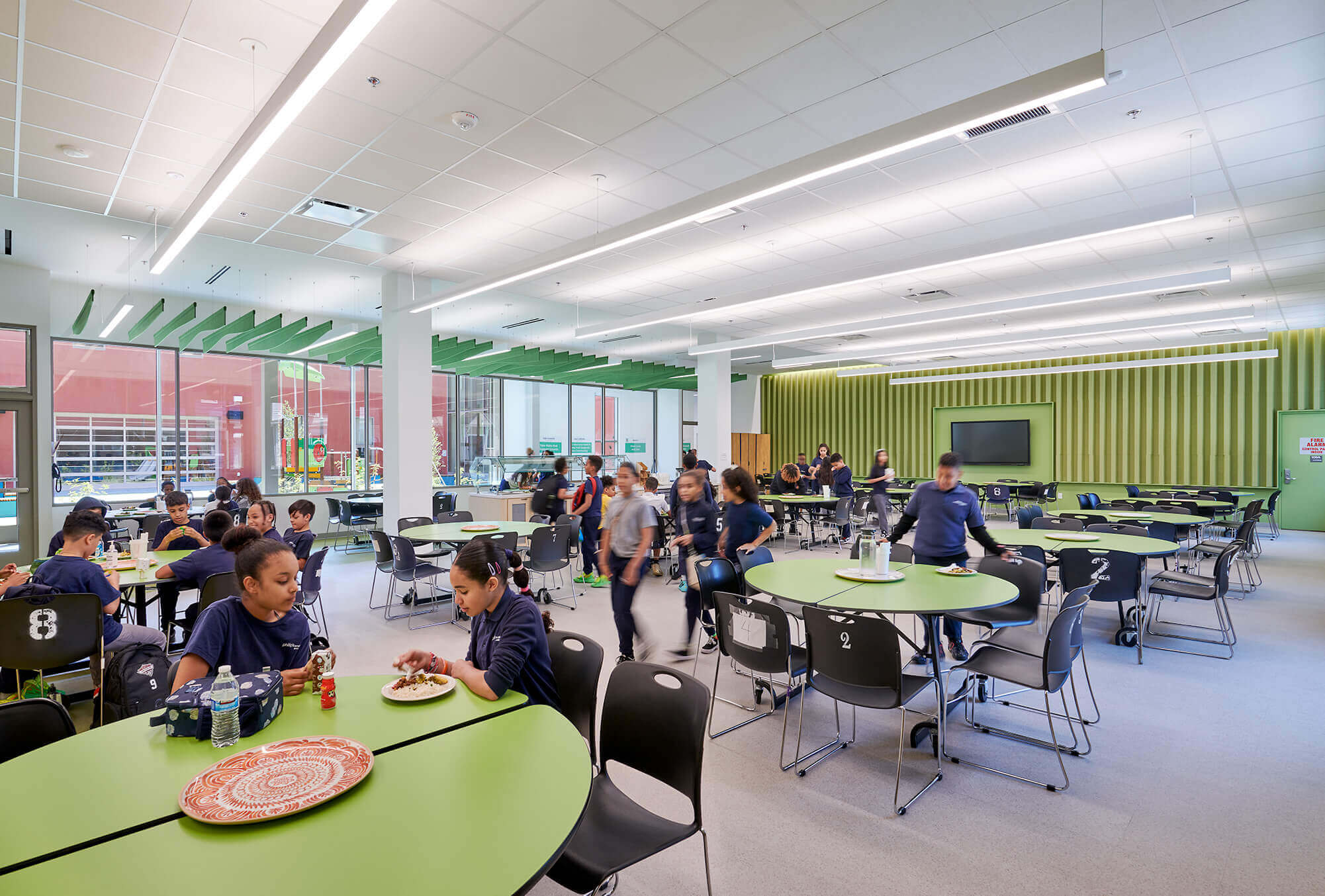 School cafeteria with students eating and walking around, featuring green tables and black chairs under bright overhead lighting.
