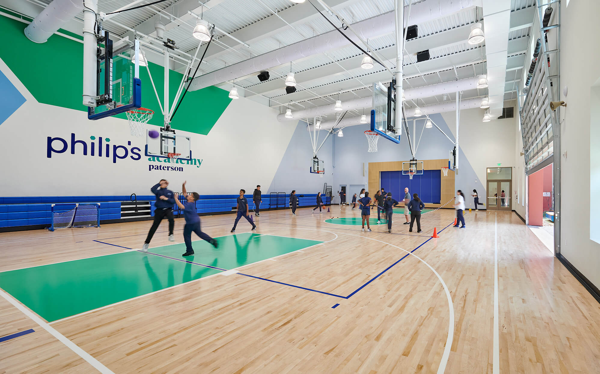 Children playing basketball and participating in activities in a spacious gymnasium with blue and green accents and 'philip's academy paterson' on the wall.
