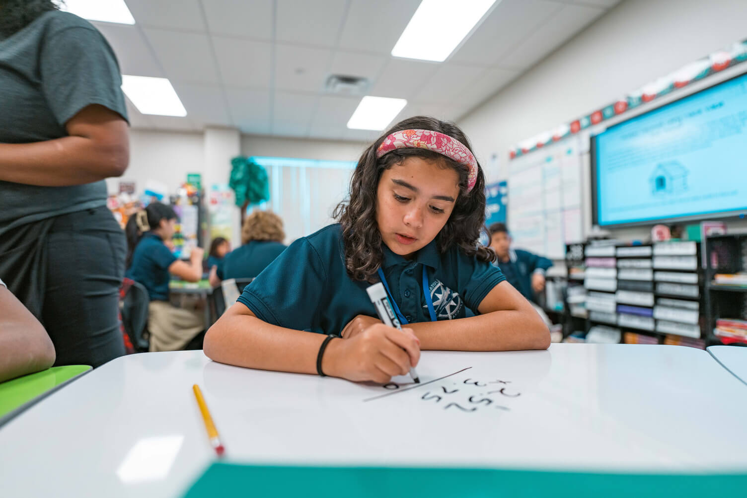 Student wearing a pink headband and school uniform writing math problems with a marker on a whiteboard table in a classroom.