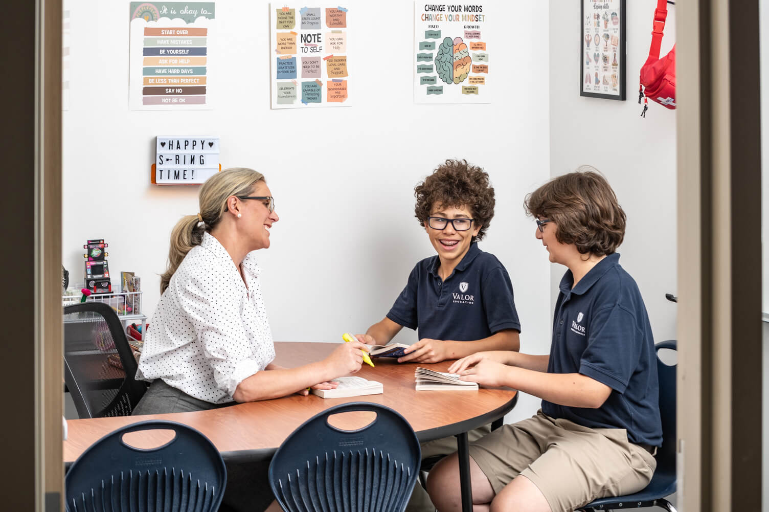 Teacher and two students in Valor Education uniforms sitting at a table smiling and reading books in a classroom with motivational posters on the wall.