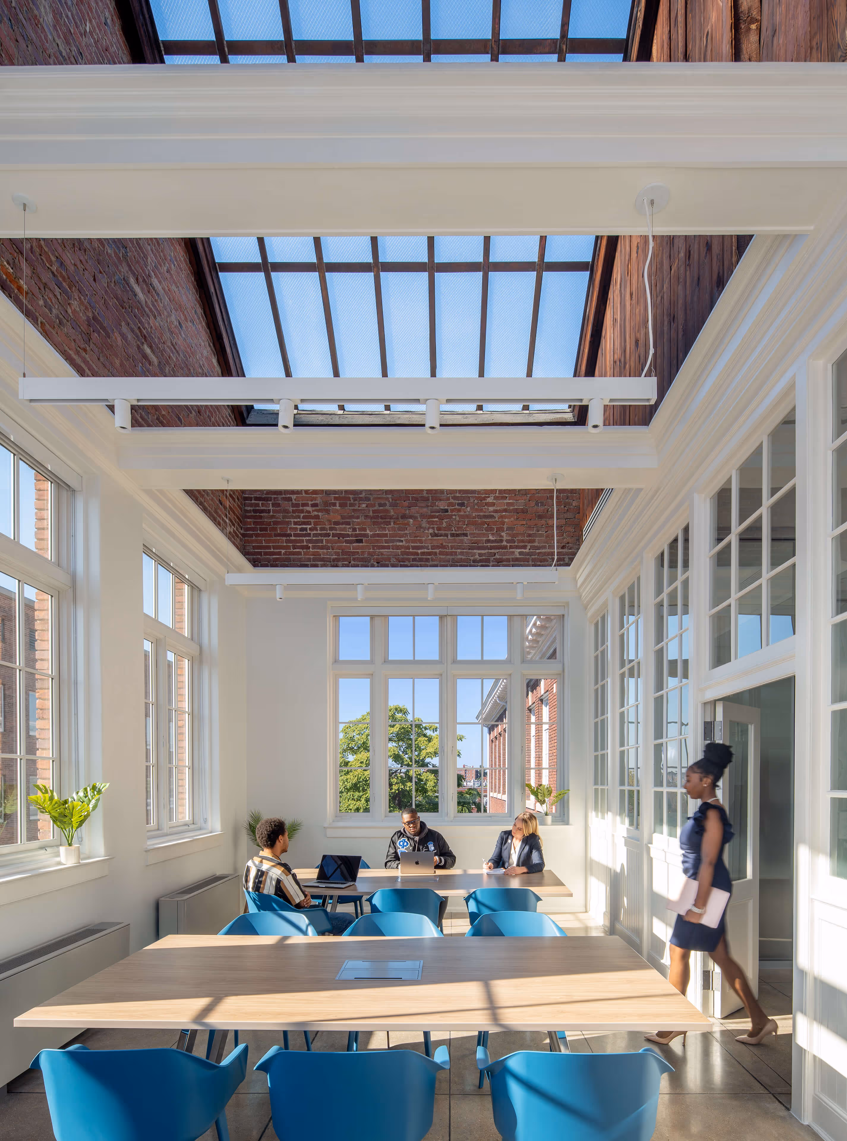 Bright modern conference room with large windows and skylight, featuring blue chairs and three people seated at a table while a woman walks by holding a folder.