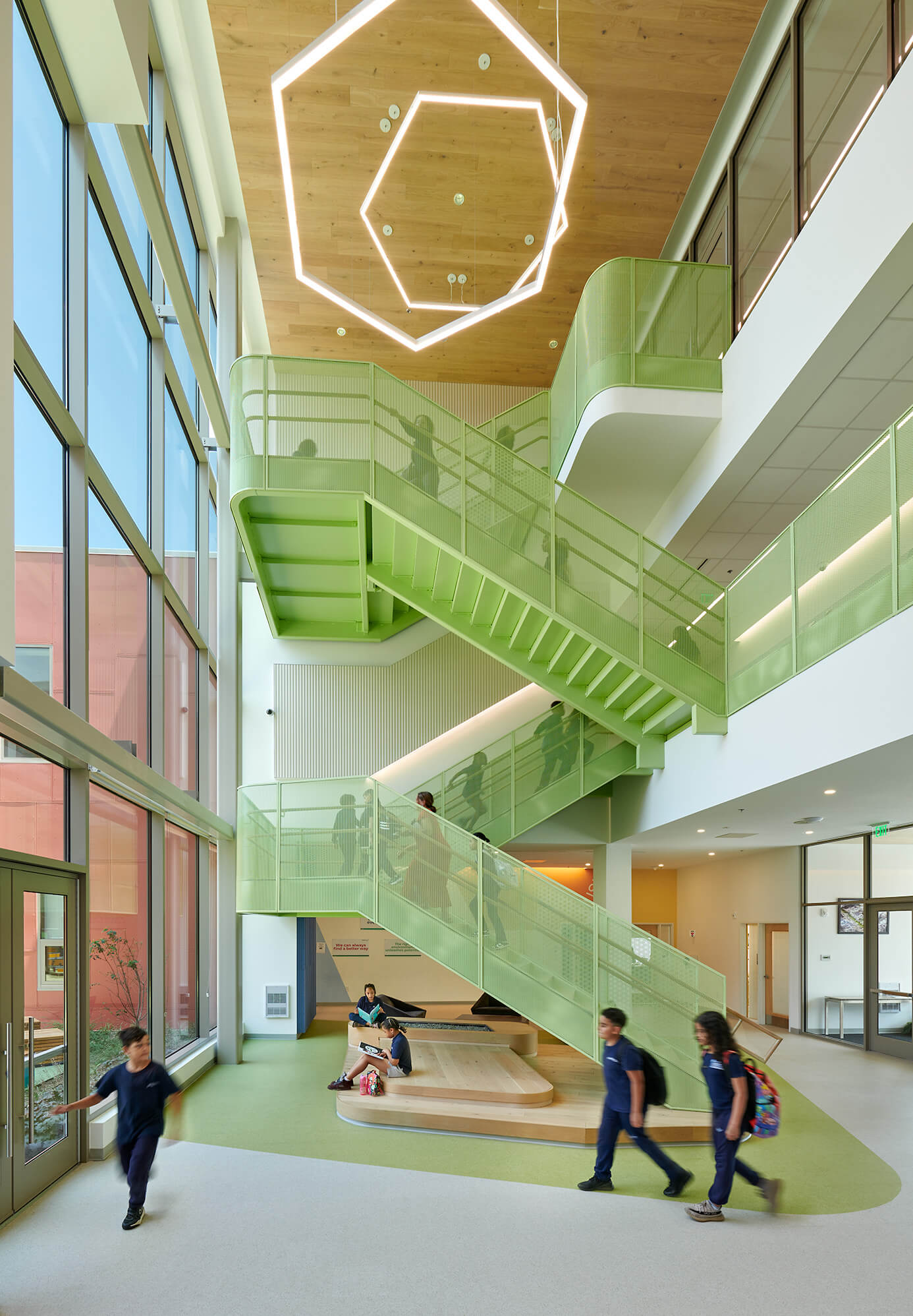 Bright modern school interior with green metal stair railings, large windows, wood ceiling fixture, and students walking and sitting.