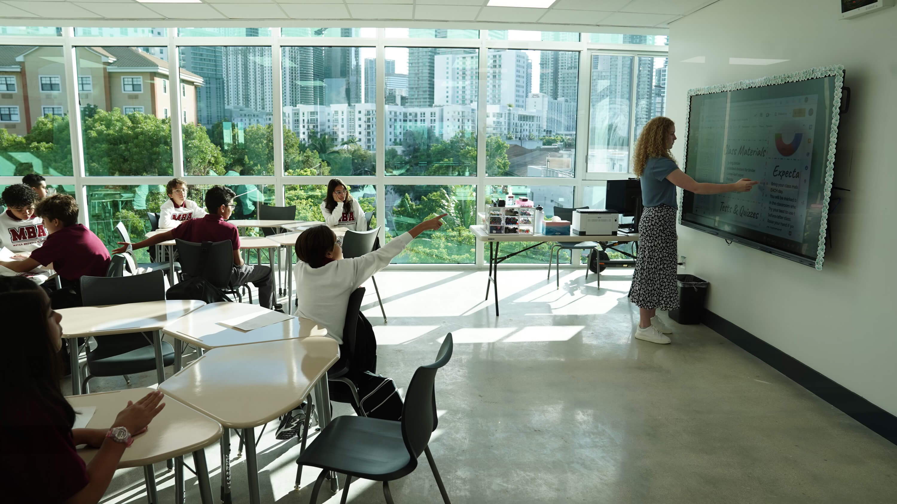 A classroom with students sitting at desks and a teacher standing at a large digital screen pointing at the lesson.