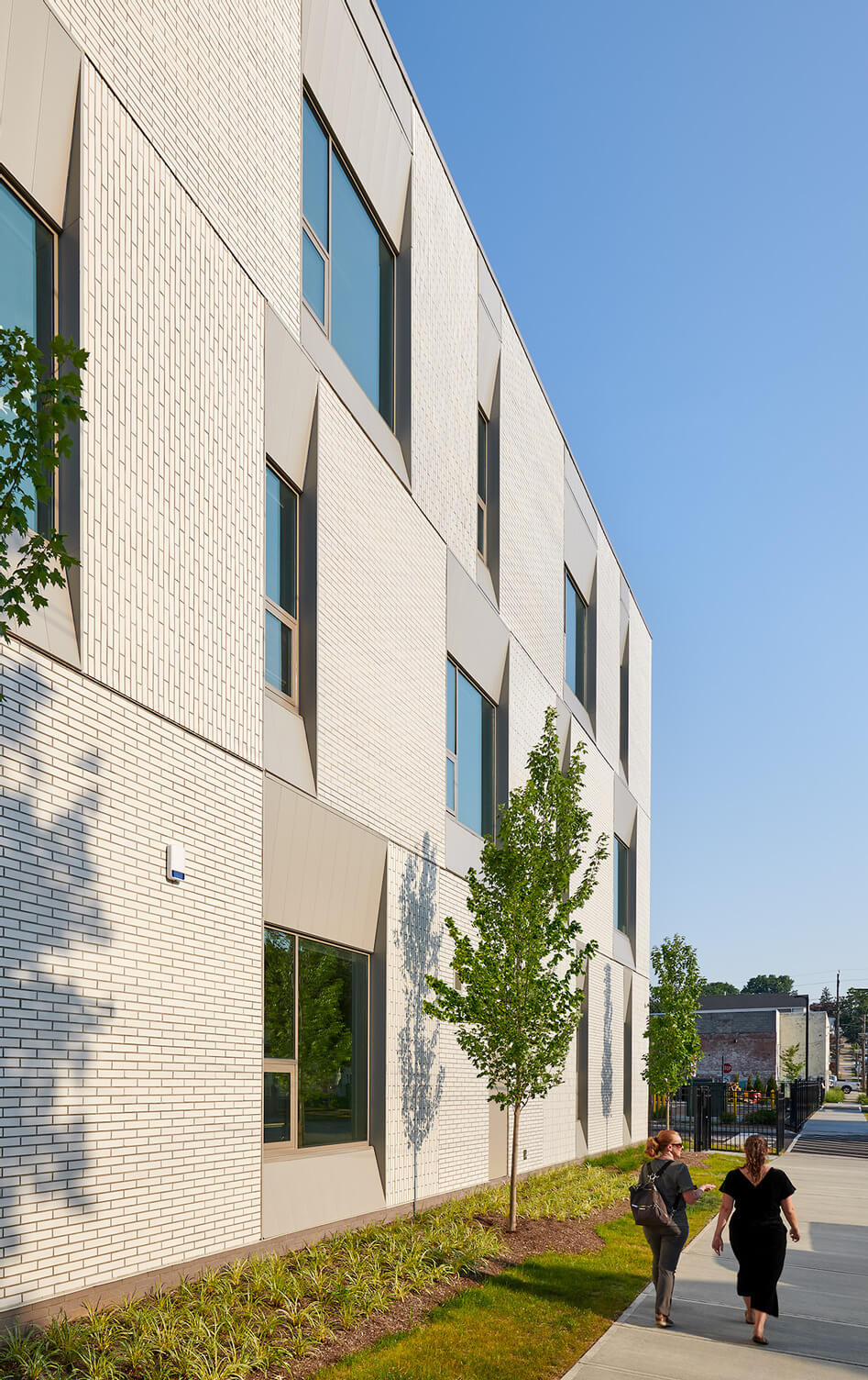 Two women walking on a sidewalk alongside a modern white brick building with tall windows and small trees lining the path under a clear blue sky.