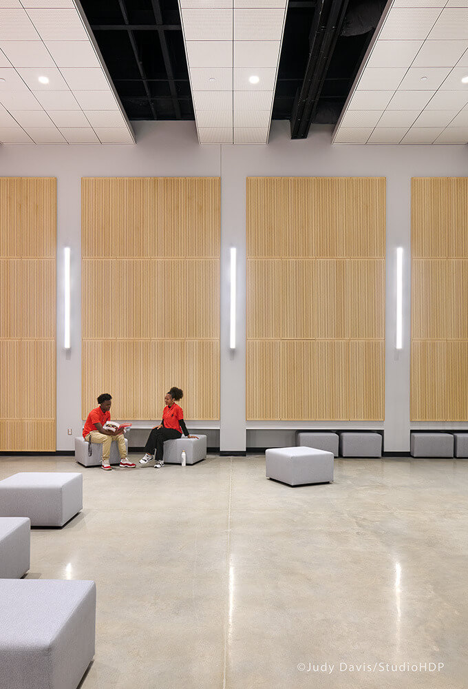 Two people in red shirts sitting on gray cube seats in a spacious room with wood panel walls and bright vertical lights.