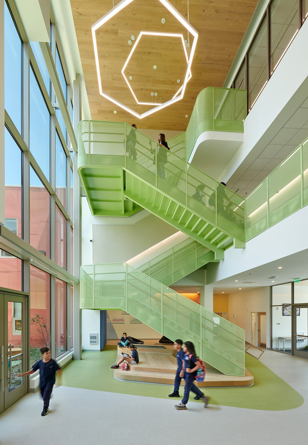 Modern school interior with large windows, pastel green staircase, students walking and sitting in common area.