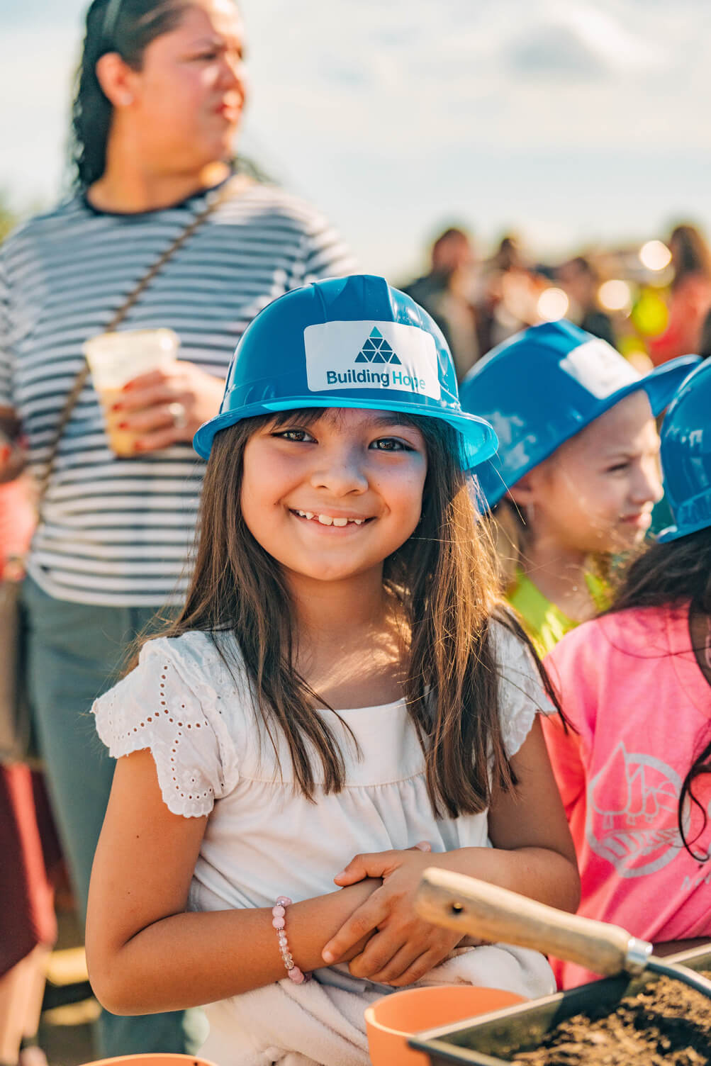 Smiling young girl wearing a blue hard hat labeled 'Building Home' at an outdoor event.