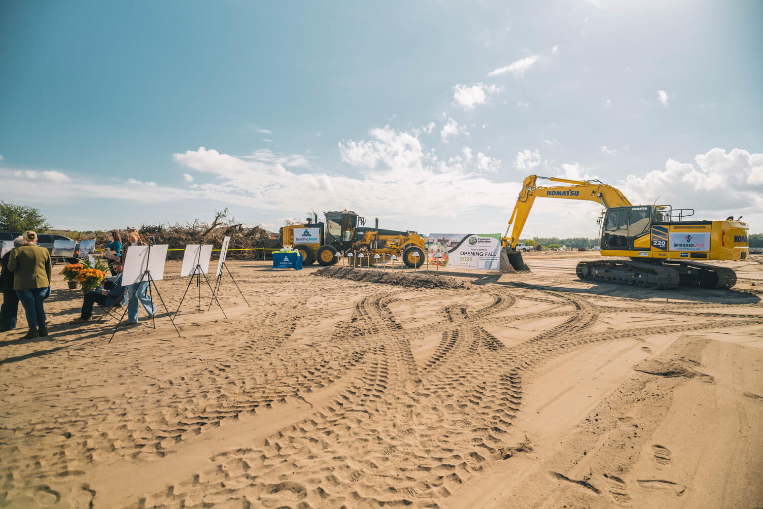 Construction site with people gathered near display boards, heavy machinery, and a banner announcing a new campus opening in fall 2026.