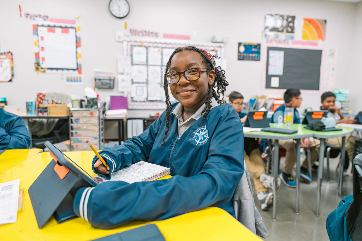 Smiling student in a blue Navigator Academy jacket writing with a pencil at a yellow table in a classroom.