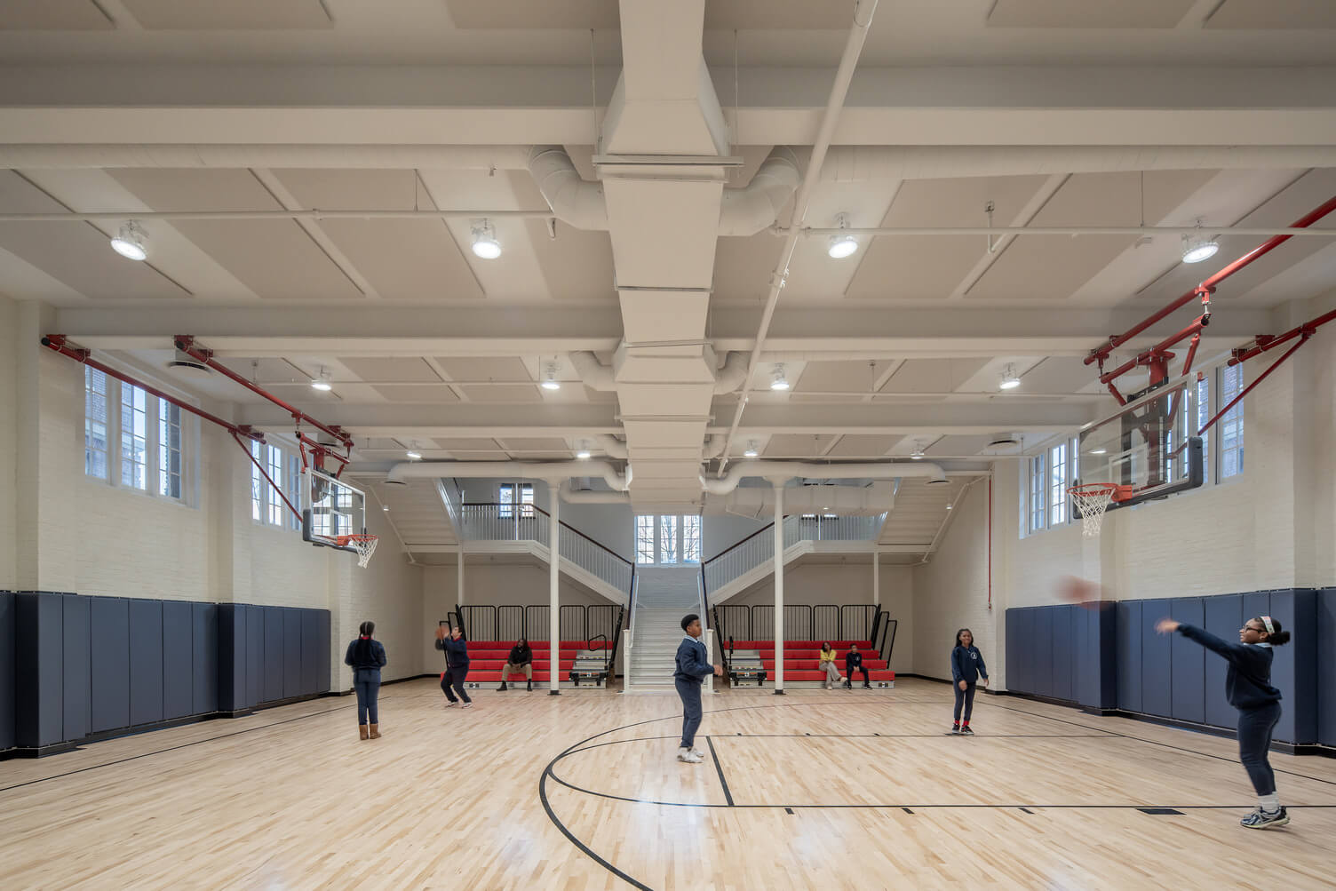 Children playing basketball in a bright indoor gym with wooden floor and red bleachers.
