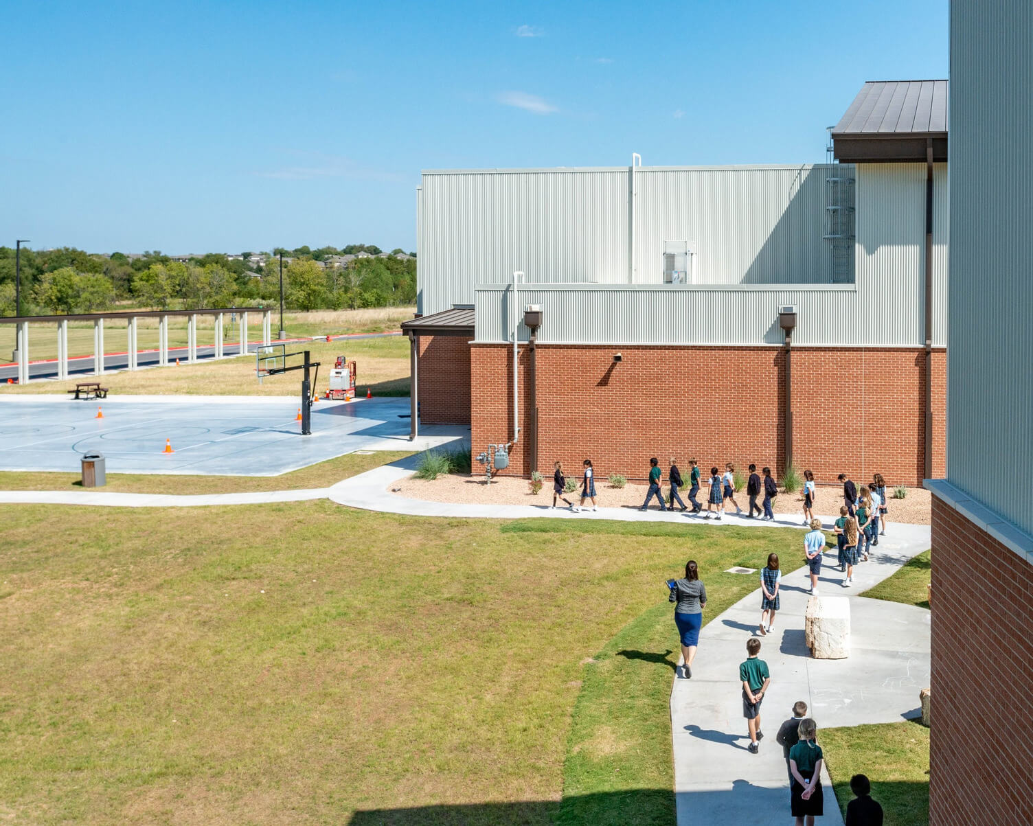 Line of students walking on a sidewalk next to a school building under a clear blue sky.