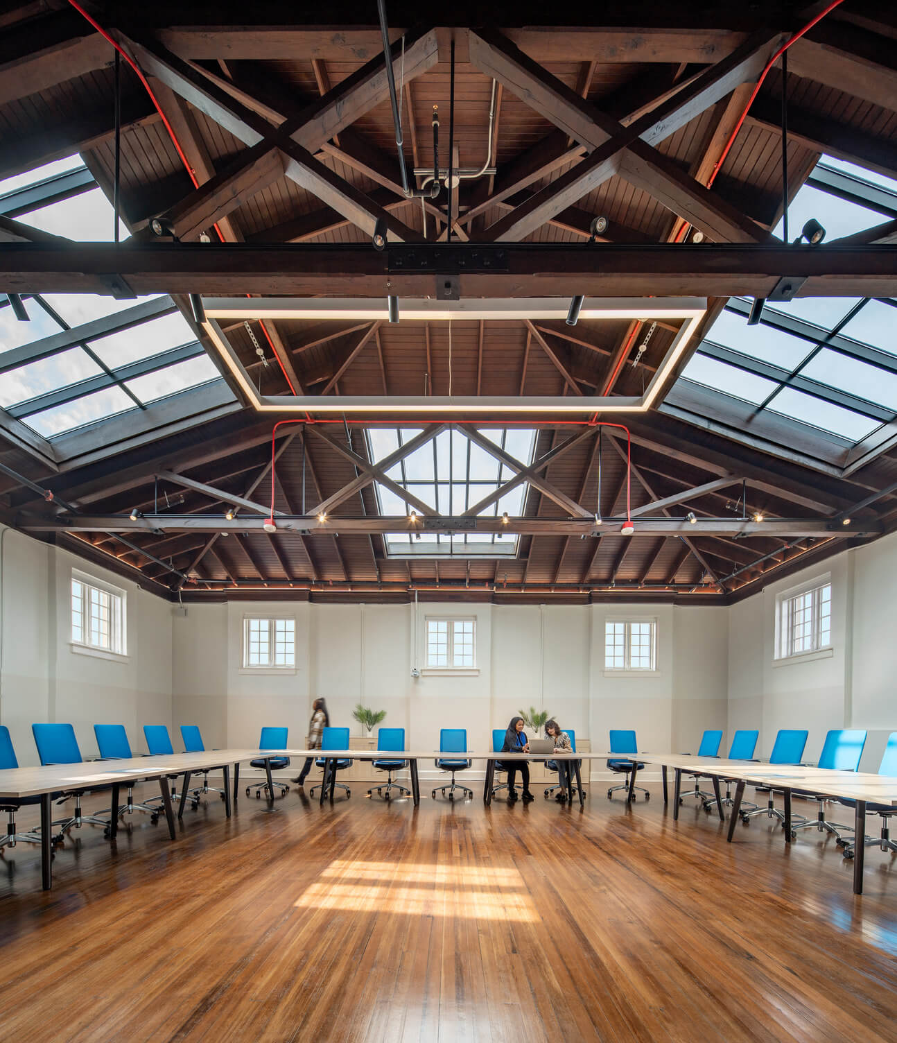 Large modern conference room with wooden floors, blue chairs around U-shaped tables, two people seated and one walking, and a wooden ceiling with skylights.