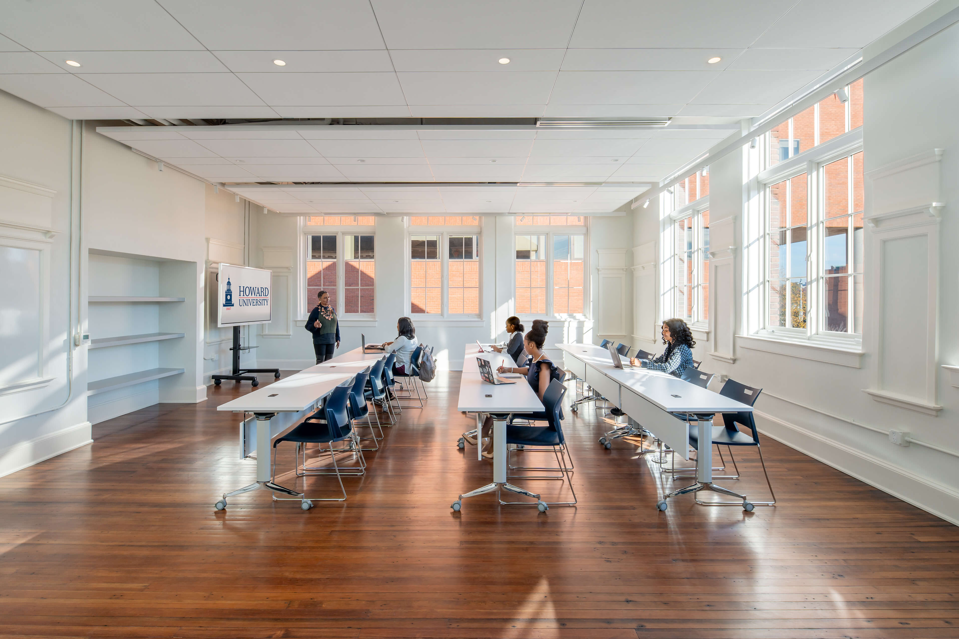 Bright classroom with large windows, a woman standing near a screen showing Howard University logo, and students seated at tables working on laptops and taking notes.
