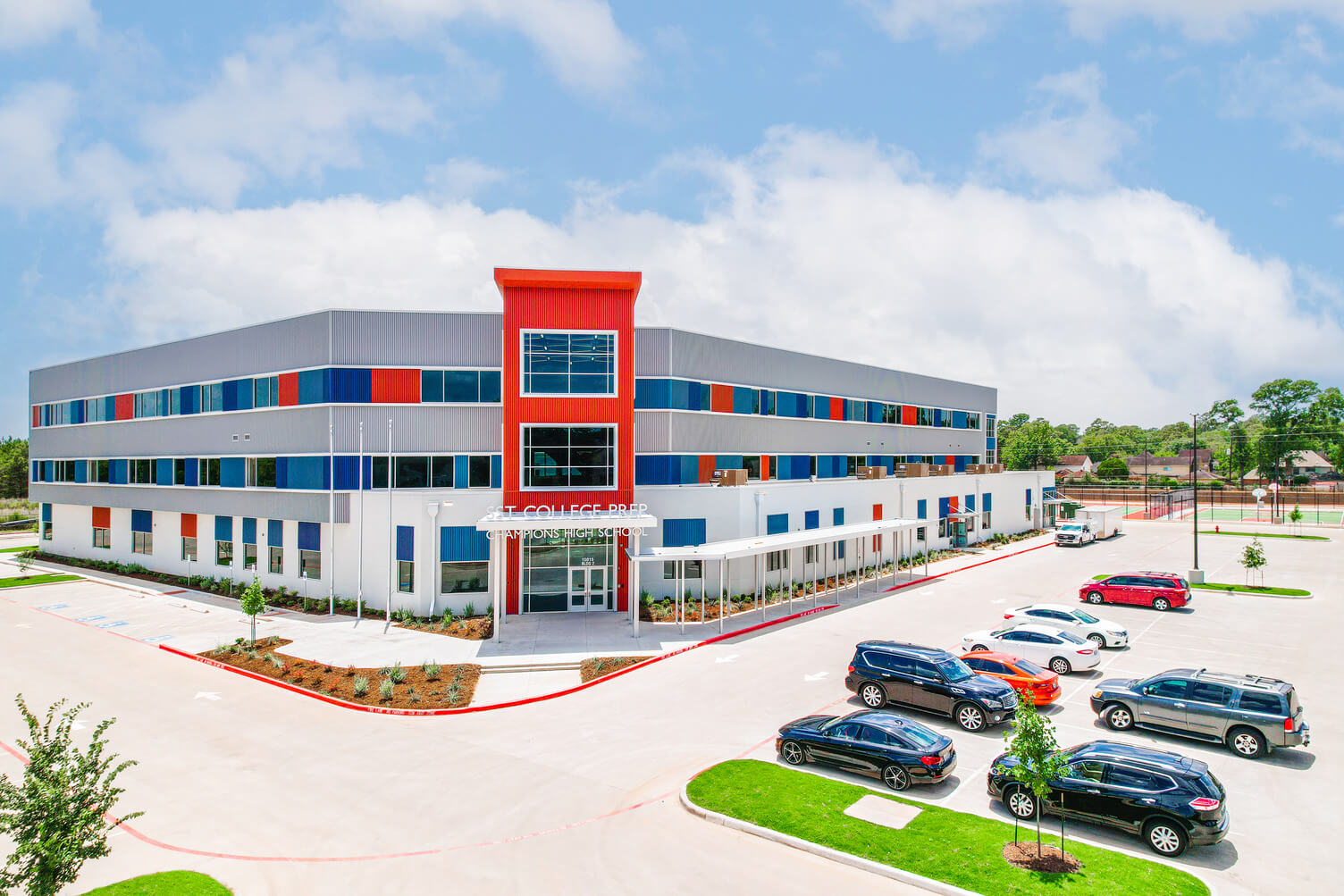 Modern three-story school building with gray, blue, and orange accents and a parking lot with cars in front.