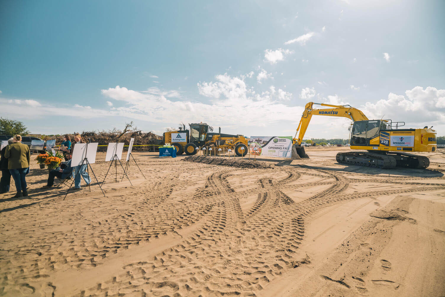 Groundbreaking event on a sunny construction site with heavy machinery, banners, and a few people gathered near display boards.