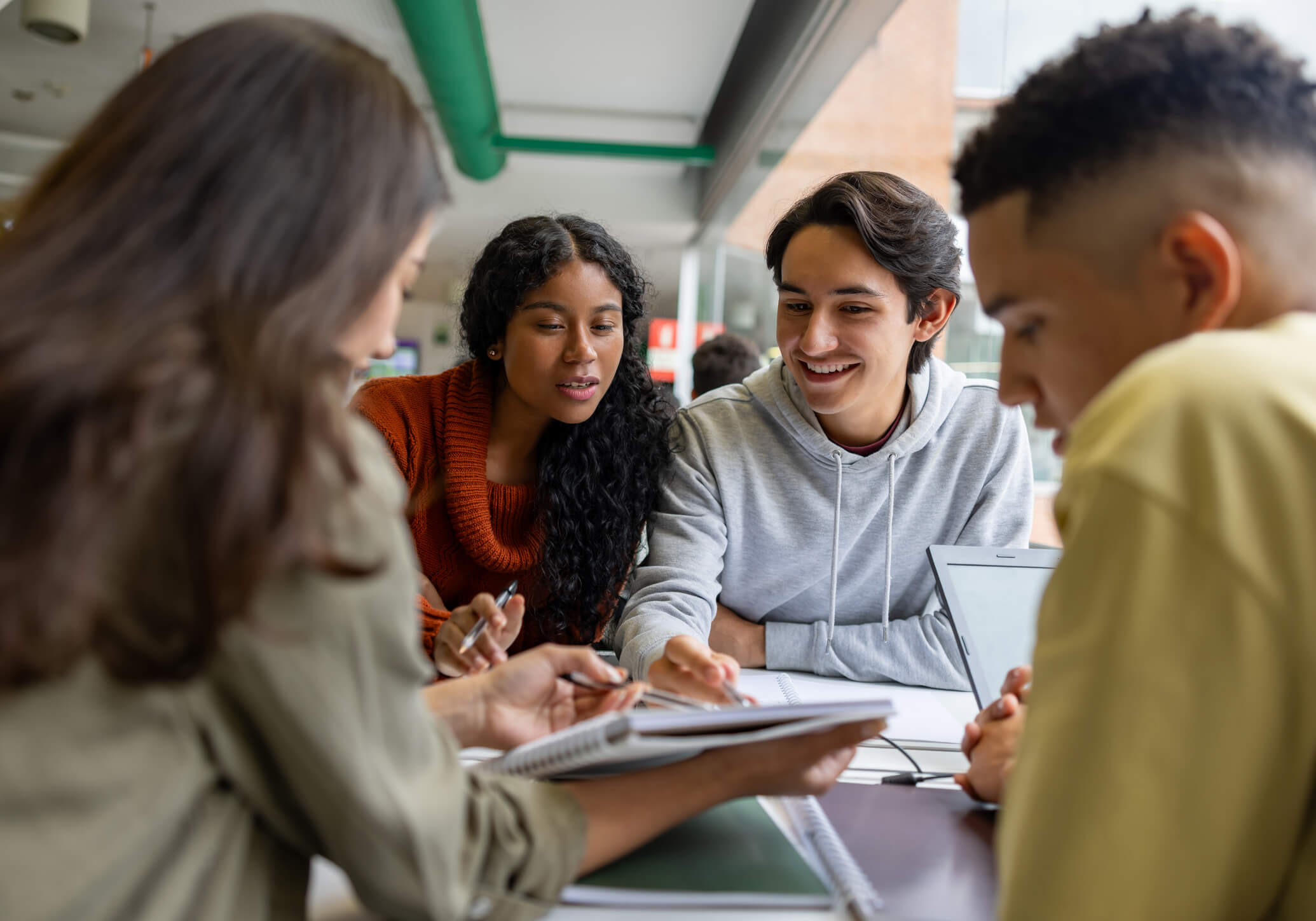 Group of young adults studying together at a table with notebooks and a laptop in a bright room.