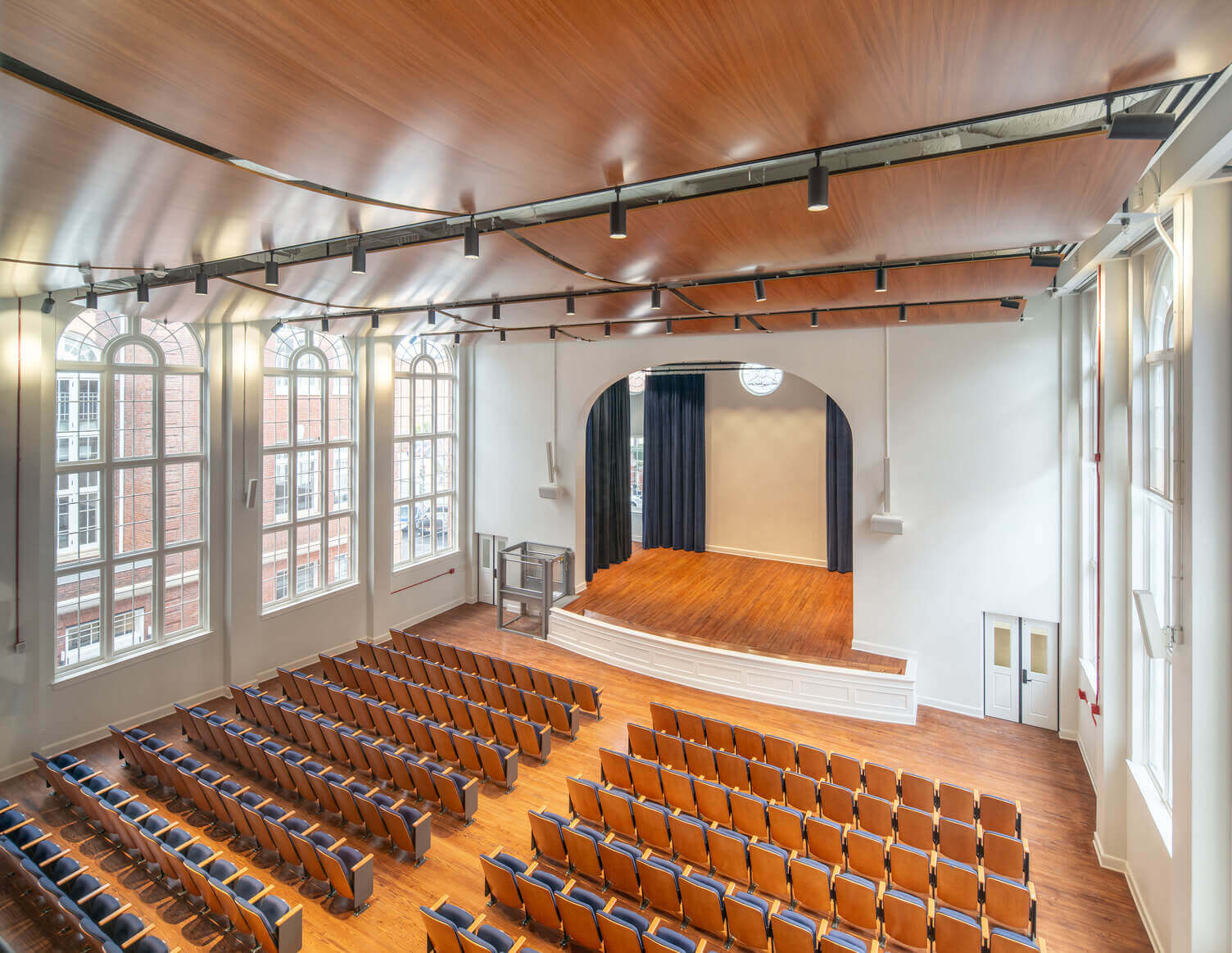 Empty auditorium with wooden floor, rows of blue and brown seats, large arched windows, and a small stage with dark curtains.