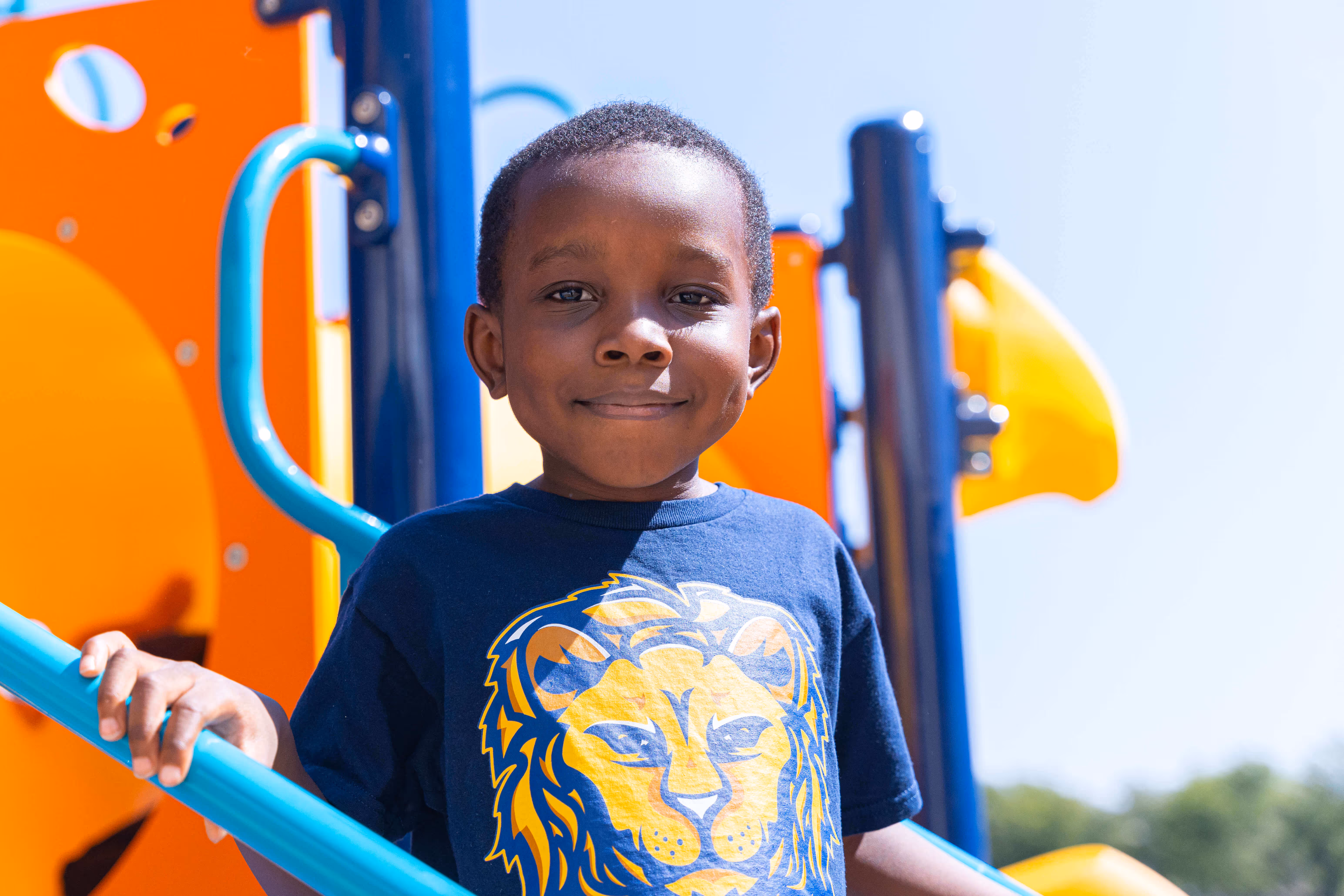 Young boy smiling and holding blue playground rails with colorful play structure in the background.