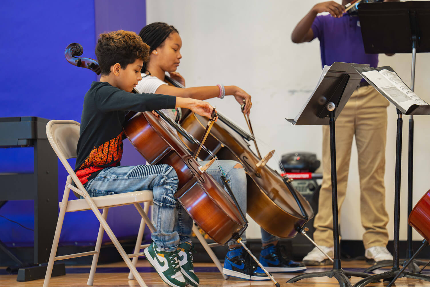 Two children seated playing cellos during a music class with a standing instructor holding a violin in the background.
