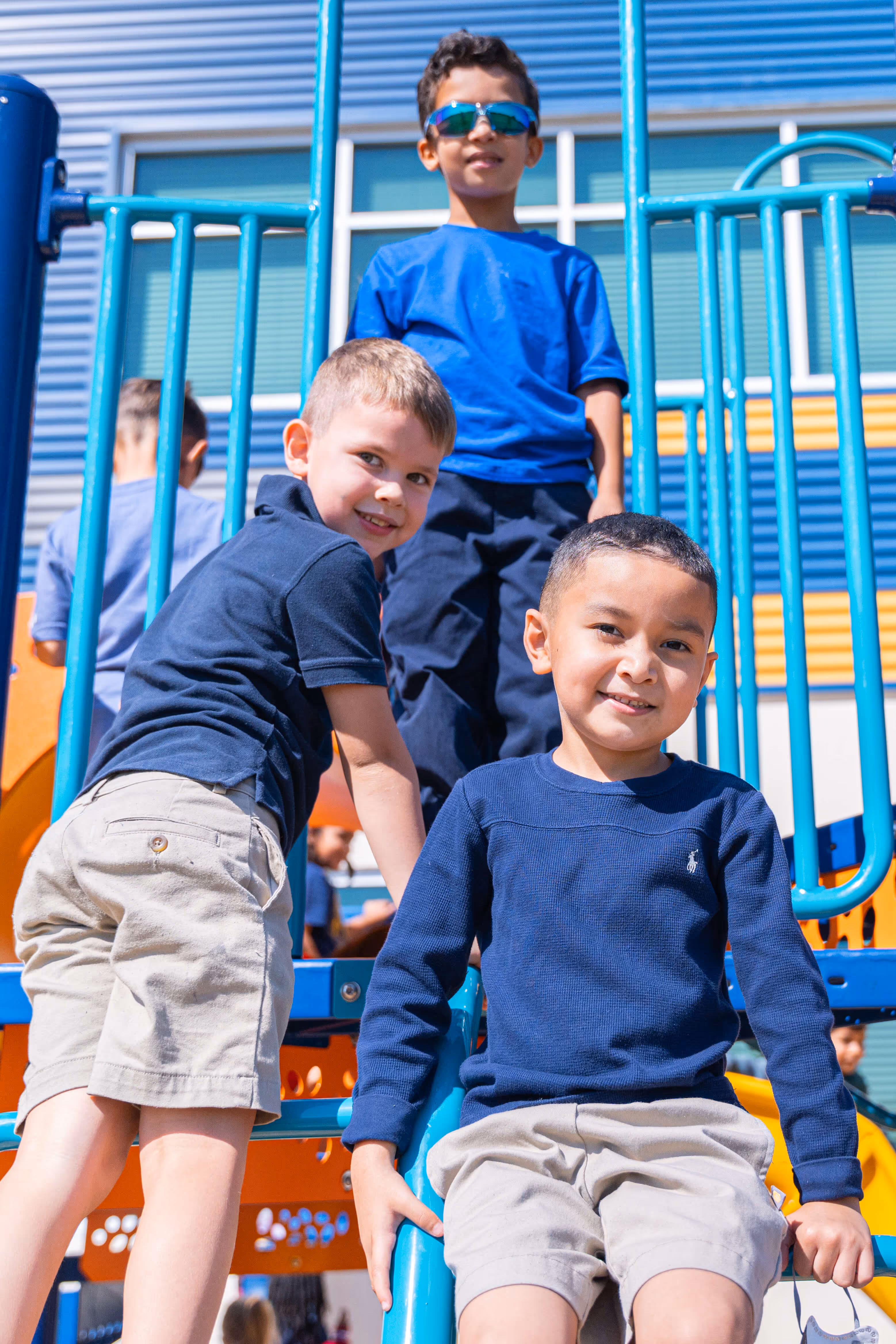 Three young boys playing on blue and orange playground equipment, smiling at the camera on a sunny day.