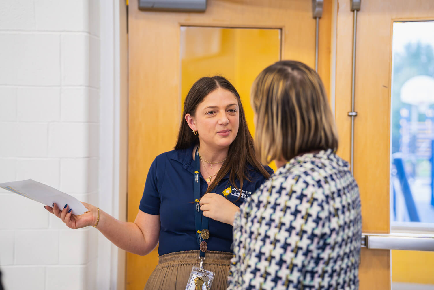 A woman in a navy blue shirt with lanyard and keys talks to another person wearing a patterned jacket near a wooden door.
