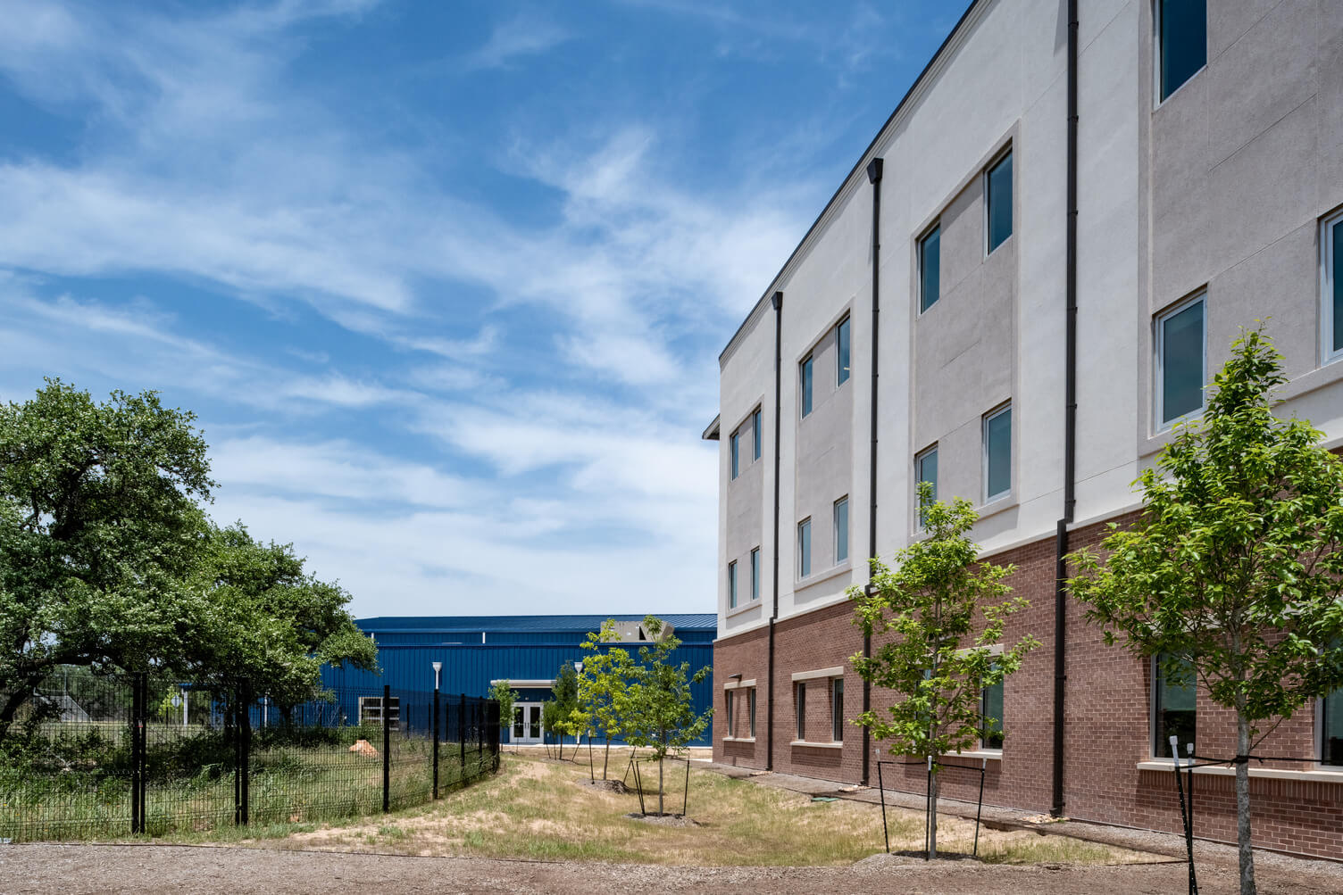 Side view of a modern three-story building with brick and light exterior walls, small trees along the path, and a blue industrial building in the background under a partly cloudy sky.